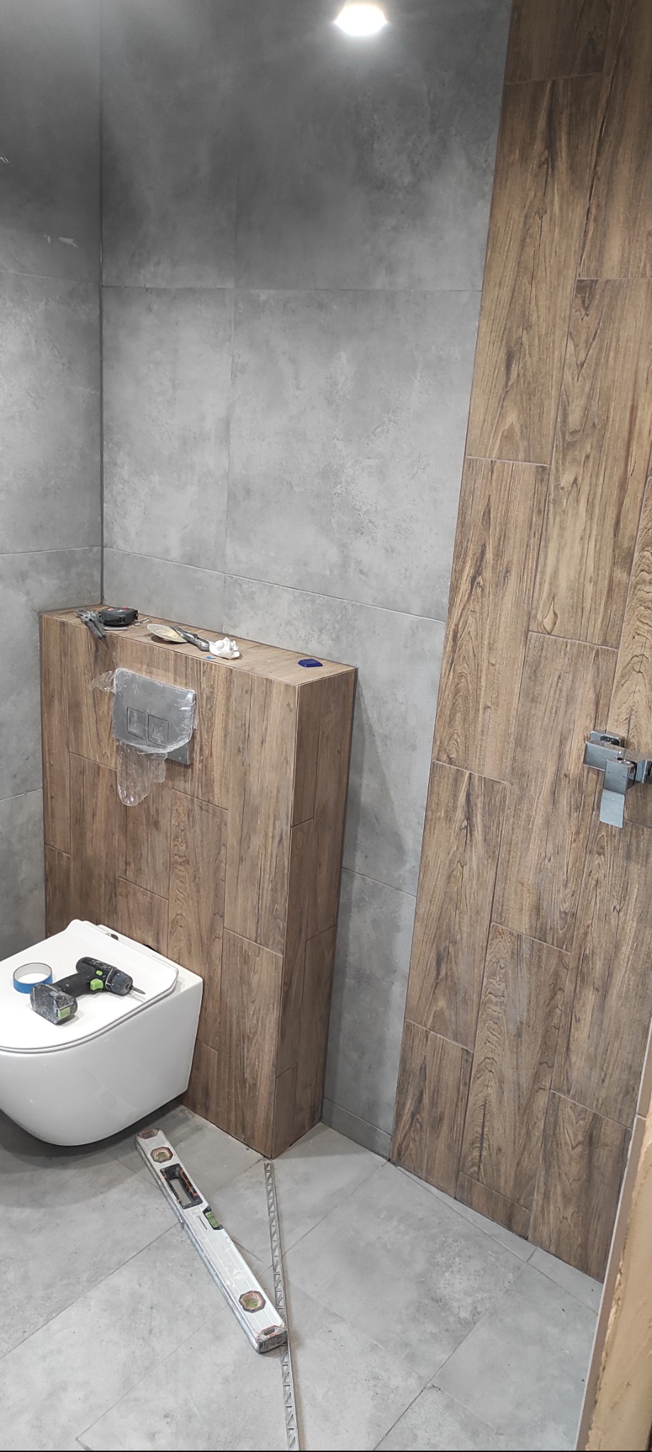 Montaż płytek imitujących drewno w łazience, widoczna toaleta podwieszana, poziomica i narzędzia glazurnicze.