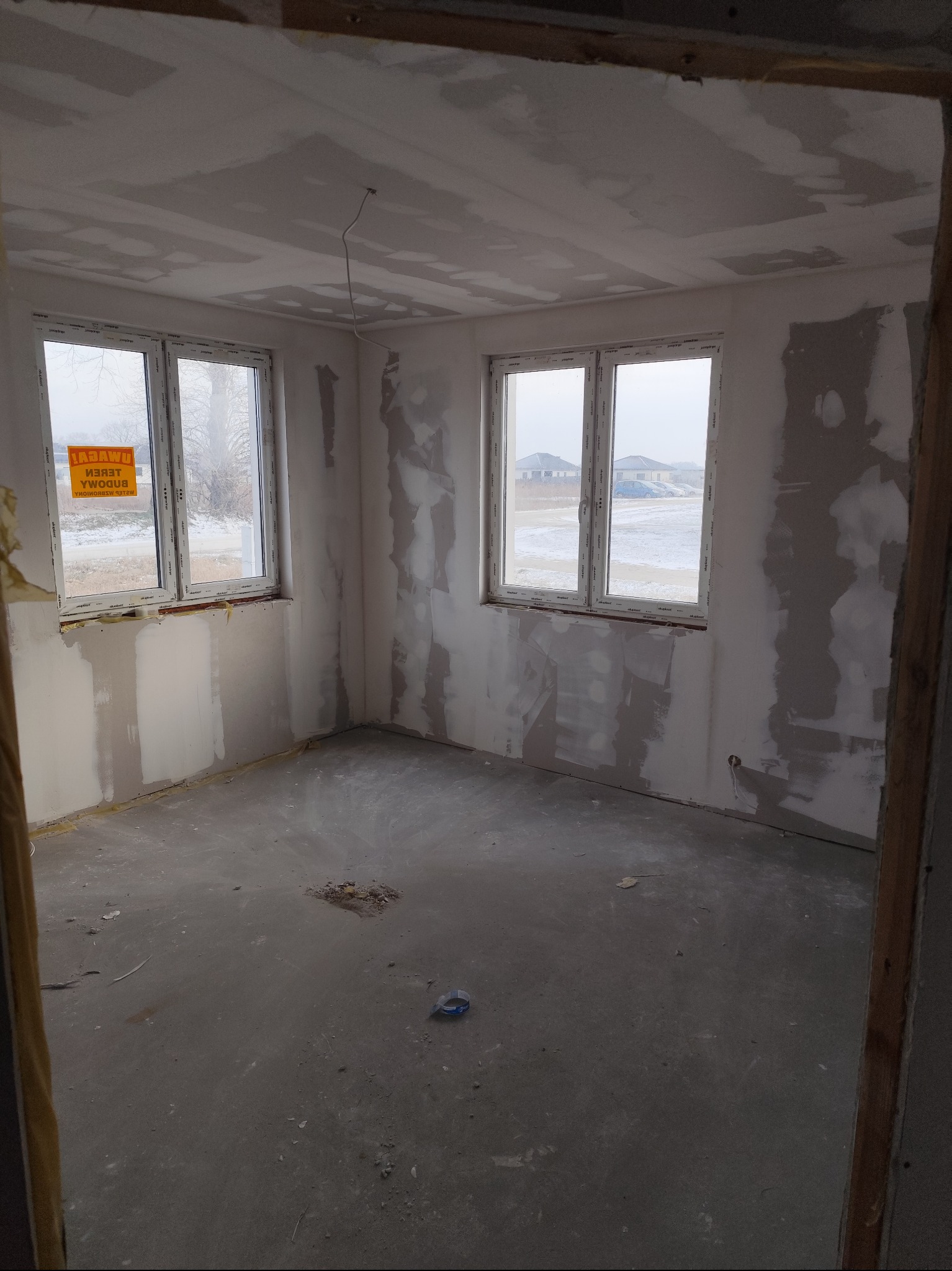 Pokój w trakcie prac wykończeniowych: ściany przygotowane do malowania, widoczne ślady szpachlowania, surowy beton na podłodze, okna z widokiem na zimowy krajobraz.