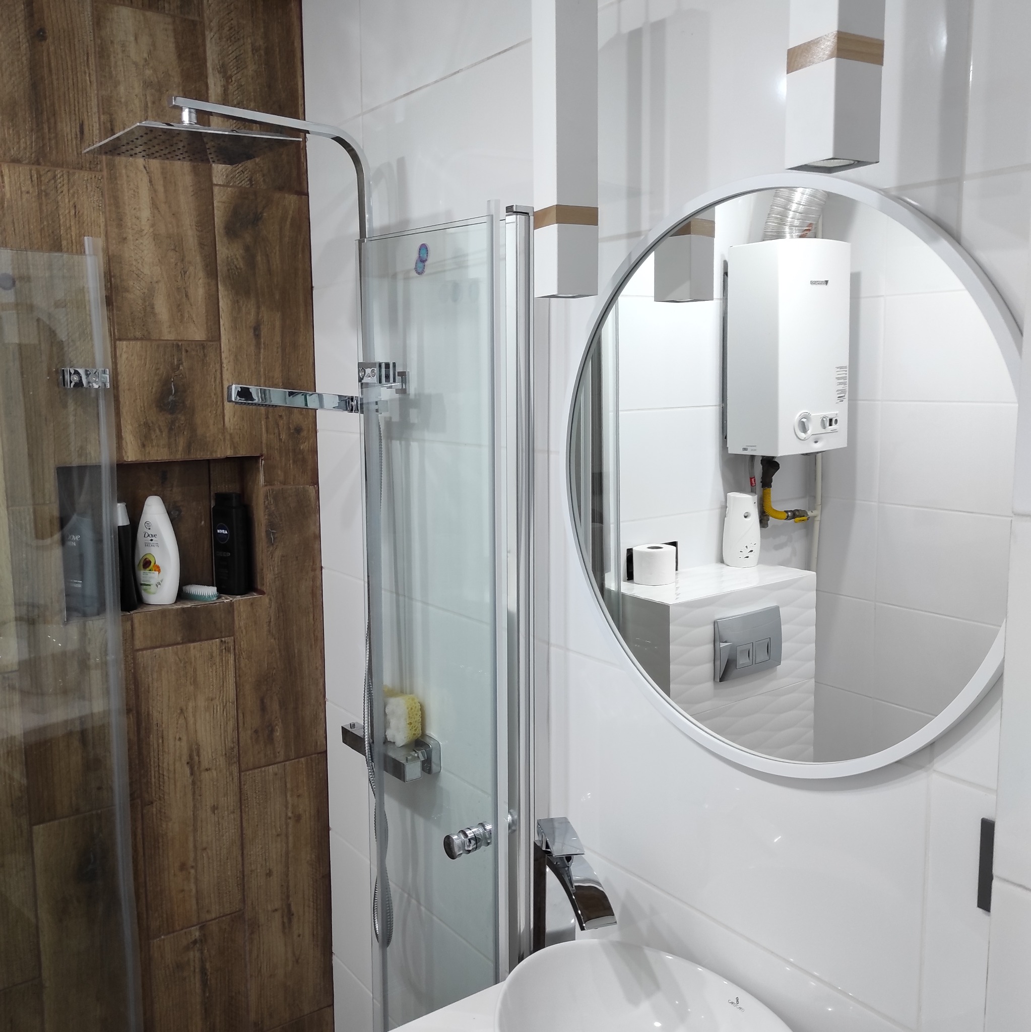 Nowoczesna łazienka z kabiną prysznicową typu walk-in, białymi płytkami i drewnianymi akcentami, widoczna armatura i okrągłe lustro.