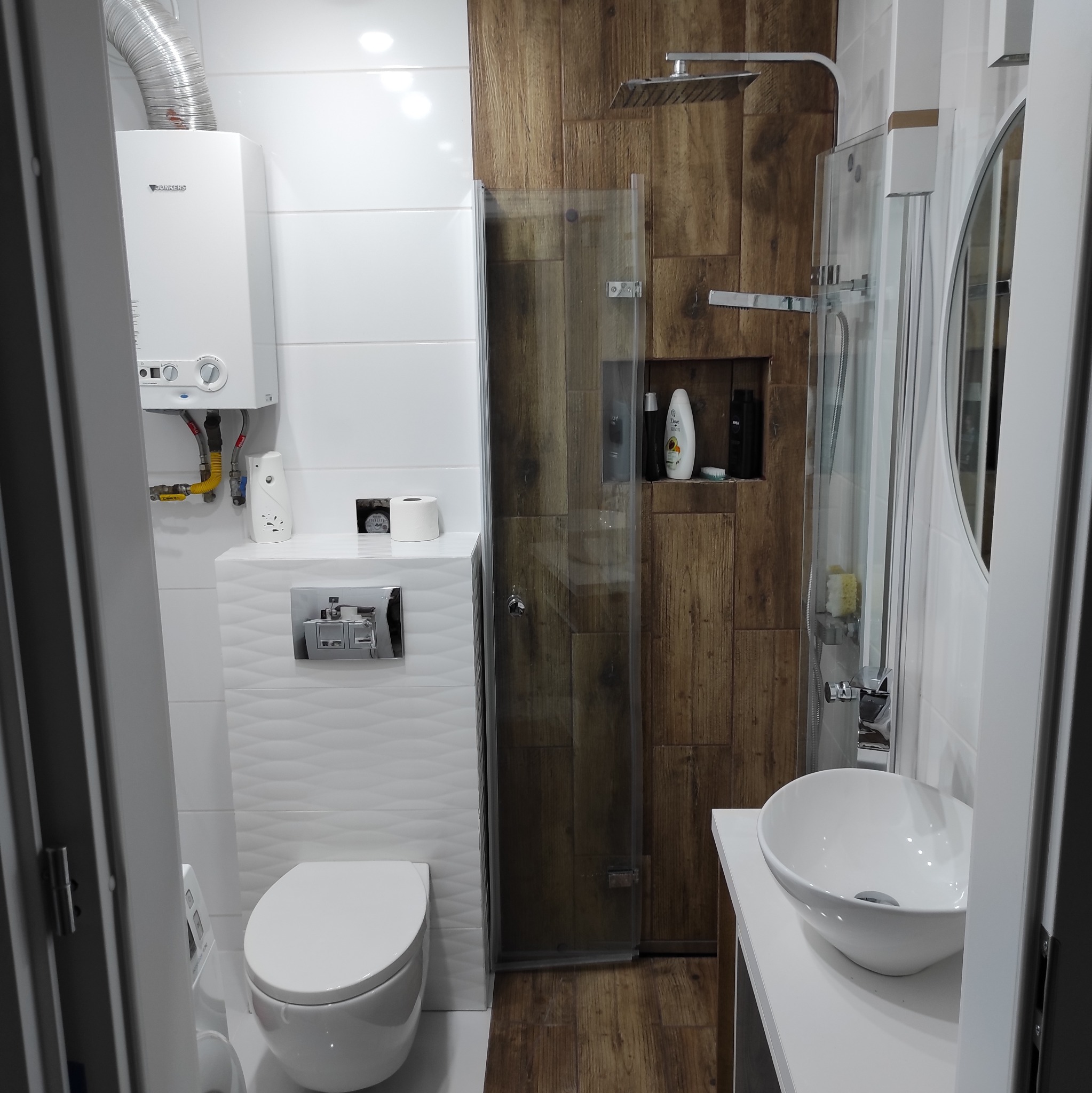 Nowoczesna łazienka z drewnianymi akcentami: kabina prysznicowa z deszczownicą, umywalka nablatowa i podwieszana toaleta. Białe płytki z fakturą i piec Junkers.