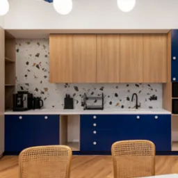 Nowoczesna kuchnia biurowa z drewnianymi szafkami, niebieskimi frontami i blatem w kolorze bieli. W tle ściana z mozaiką. Dwa krzesła z rattanu przy stole.