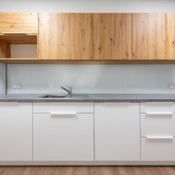 Navo Sp. Z o.o. - Nowoczesna kuchnia: białe szafki dolne z szarym blatem, drewniane szafki górne. Minimalistyczny design, widoczny fragment lodówki i drewniana podłoga.