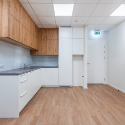 Navo Sp. Z o.o. - Nowoczesna kuchnia z drewnianymi szafkami wiszącymi i białymi szafkami dolnymi, szarym blatem oraz jasną podłogą. Widoczny fragment korytarza z białymi ścianami i drzwiami.