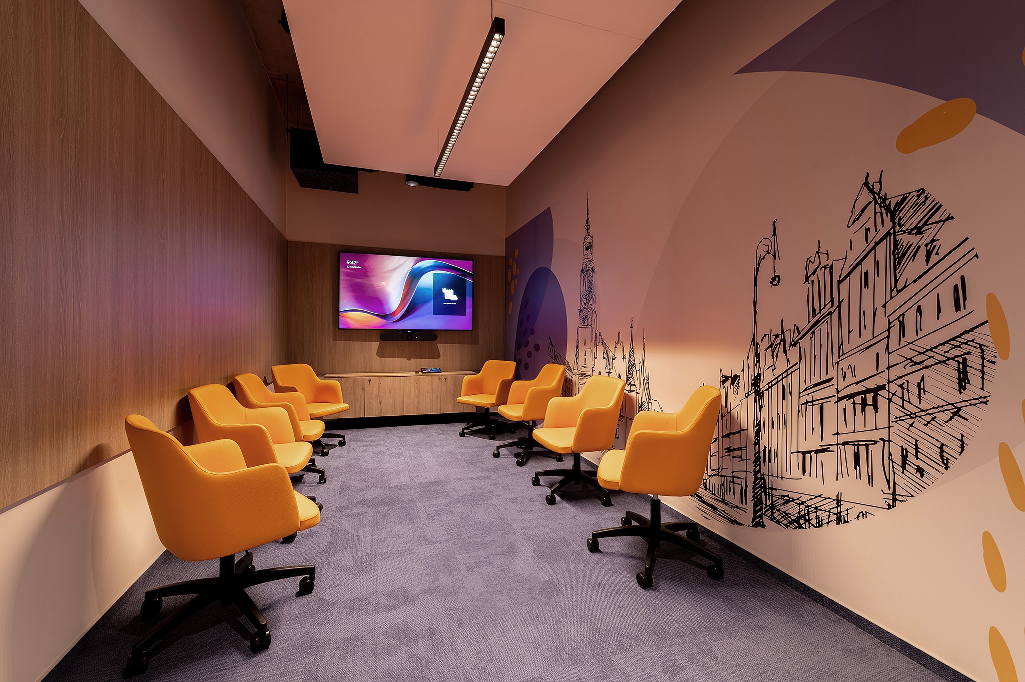 Nowoczesna sala konferencyjna z pomarańczowymi krzesłami, dużym ekranem i unikalną grafiką na ścianie przedstawiającą architekturę miejską. Jasne oświetlenie.