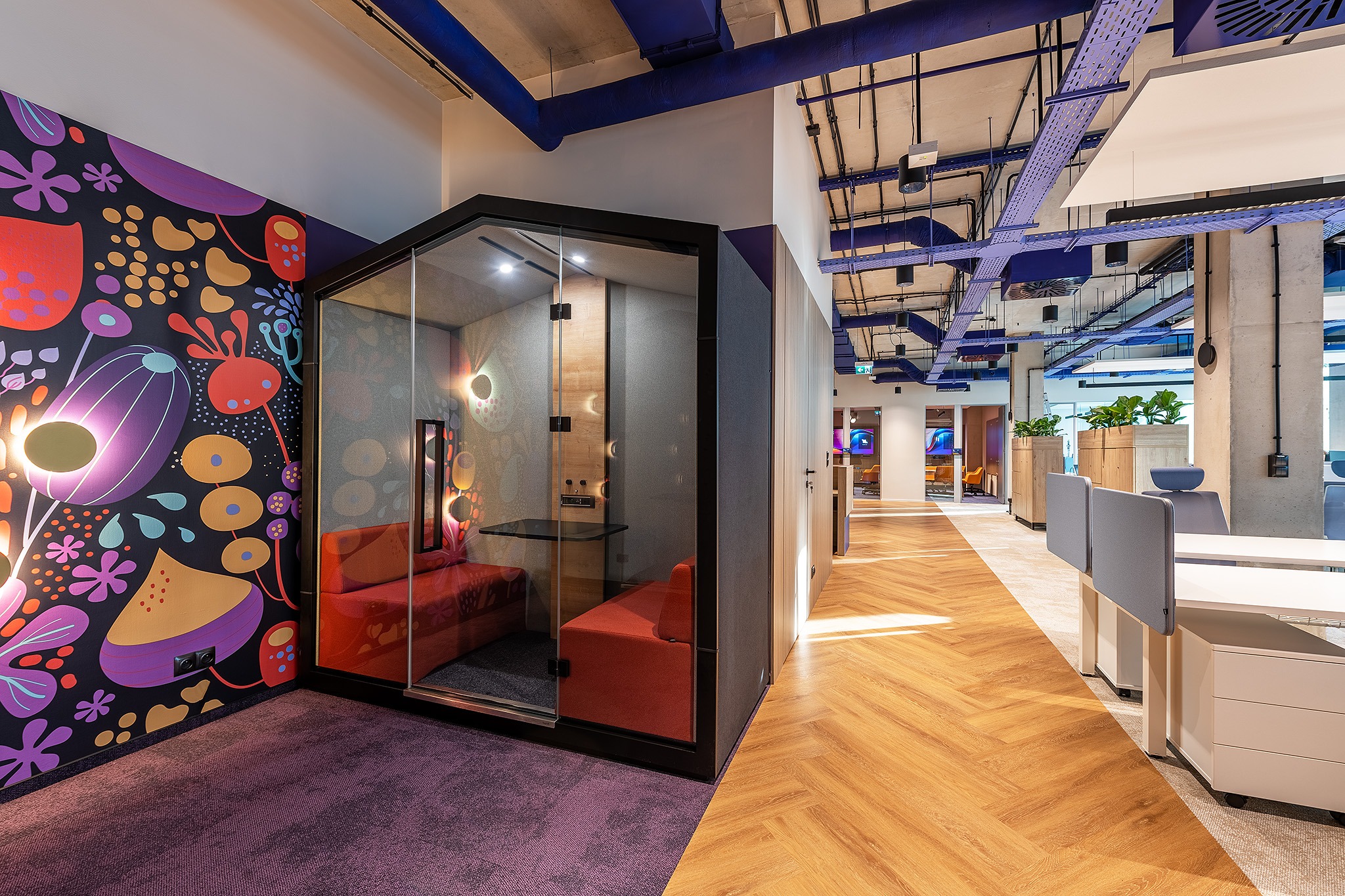 Nowoczesne biuro z kabiną akustyczną, wzorzystą tapetą i parkietem w jodełkę. Widoczne instalacje pod sufitem pomalowane na niebiesko, dodające koloru przestrzeni.