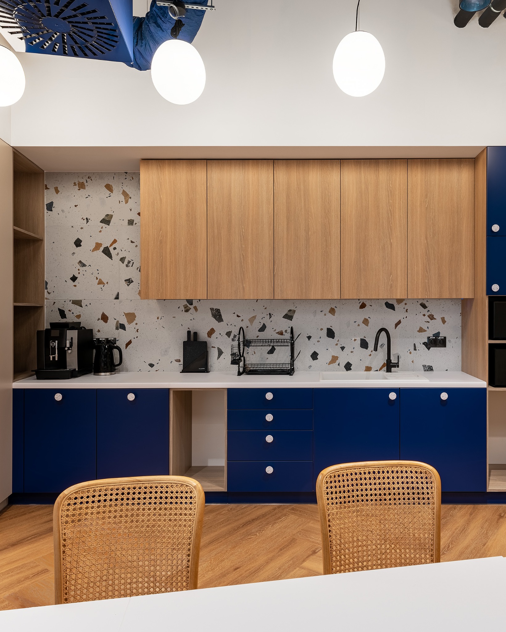 Nowoczesna kuchnia biurowa z drewnianymi szafkami, niebieskimi frontami i blatem w kolorze bieli. W tle ściana z mozaiką. Dwa krzesła z rattanu przy stole.