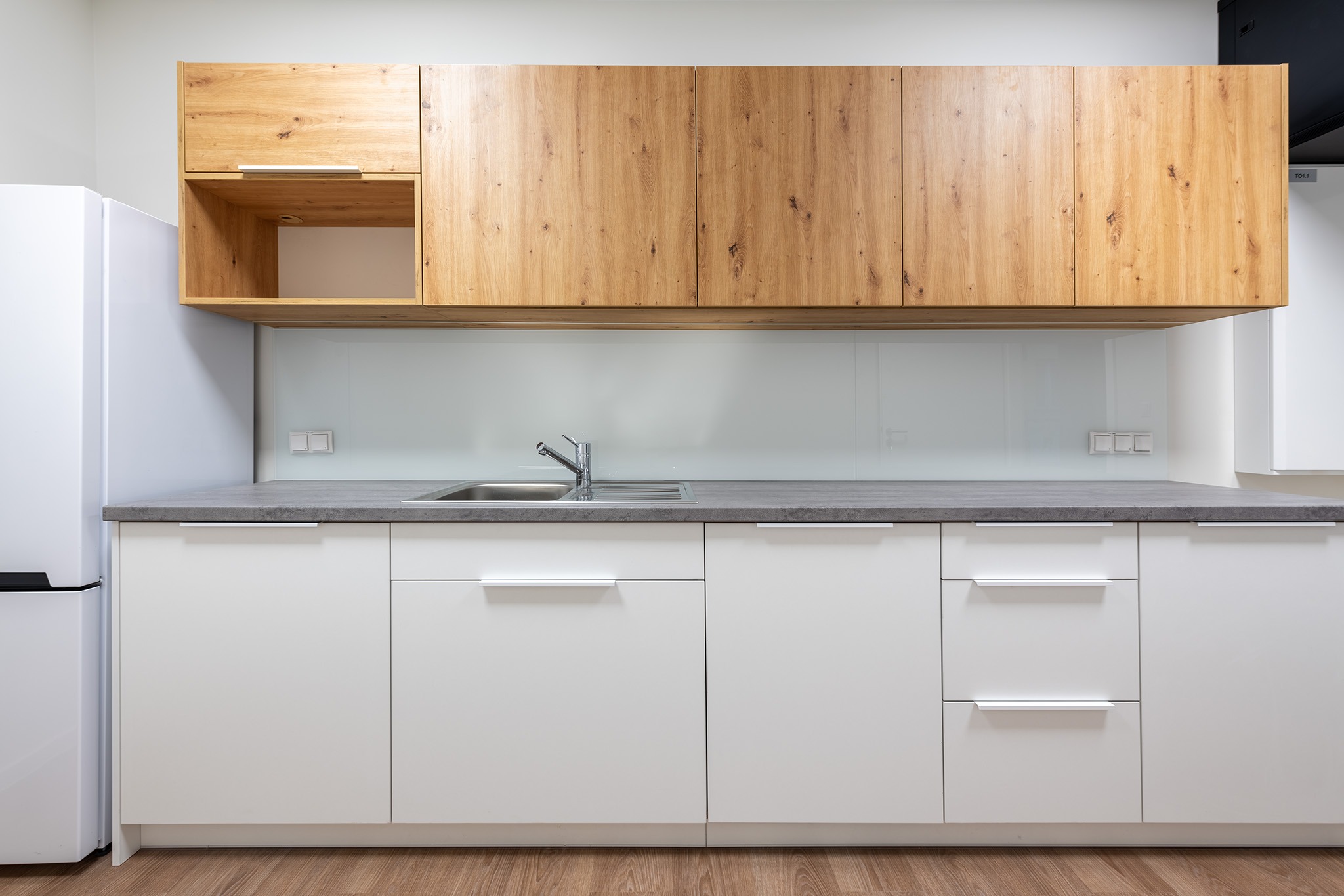 Nowoczesna kuchnia: białe szafki dolne z szarym blatem, drewniane szafki górne. Minimalistyczny design, widoczny fragment lodówki i drewniana podłoga.