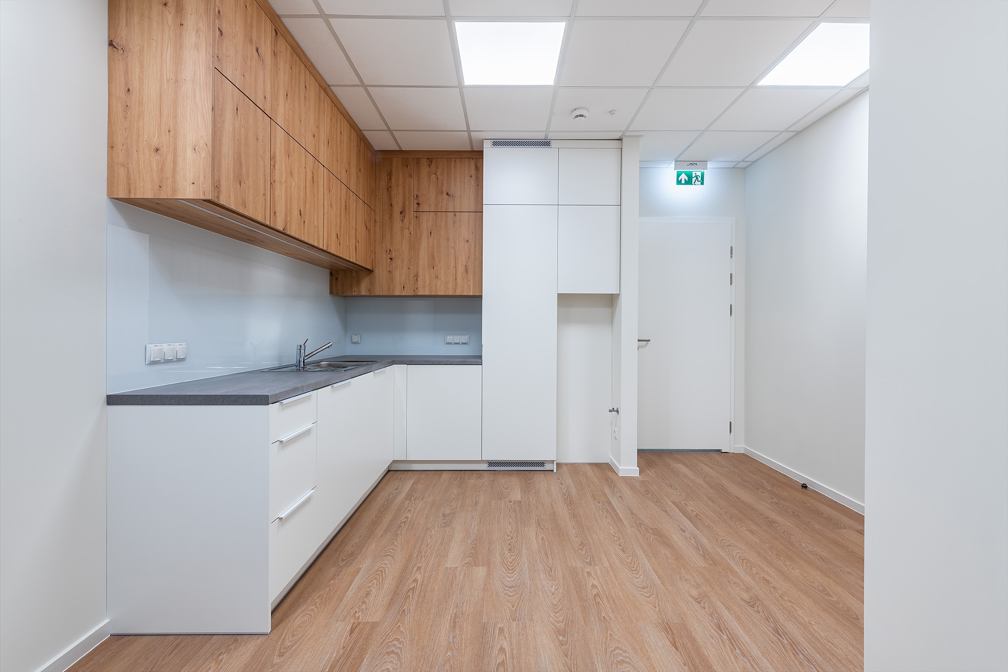 Nowoczesna kuchnia z drewnianymi szafkami wiszącymi i białymi szafkami dolnymi, szarym blatem oraz jasną podłogą. Widoczny fragment korytarza z białymi ścianami i drzwiami.