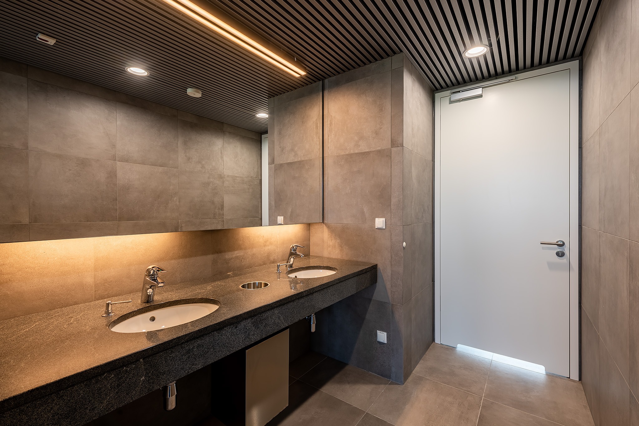 Nowoczesna łazienka biurowa z dwoma umywalkami na granitowym blacie, szare płytki i podświetlany sufit lamelowy. Białe drzwi po prawej stronie.