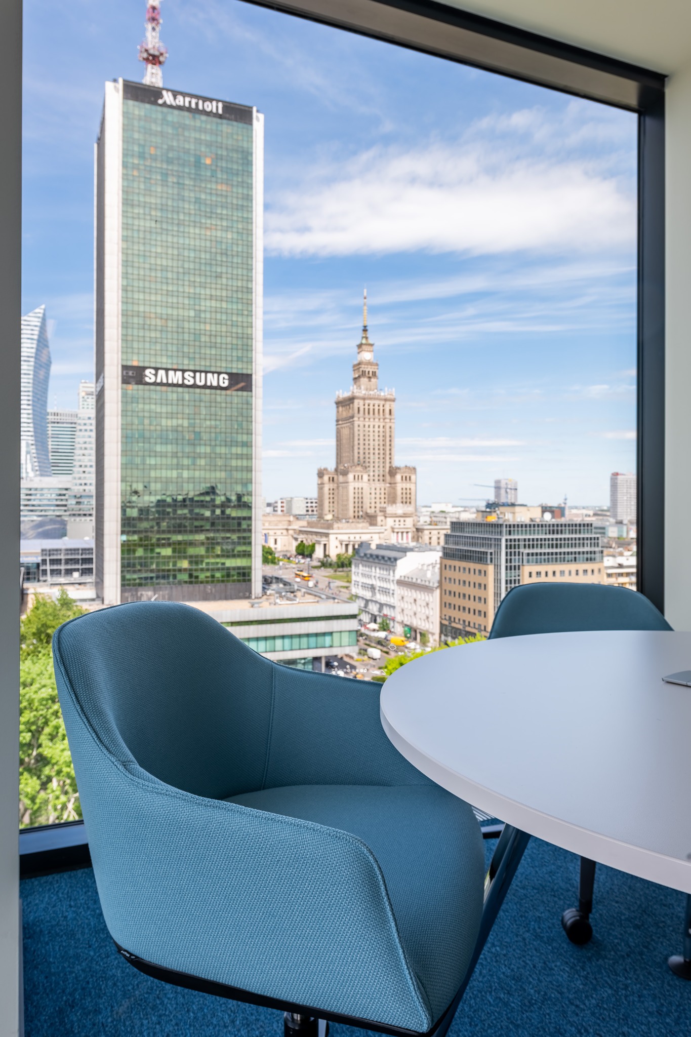 Nowoczesne biuro z widokiem na Warszawę: błękitne krzesła i okrągły stół na tle Pałacu Kultury i wieżowców Marriotta i Samsunga.