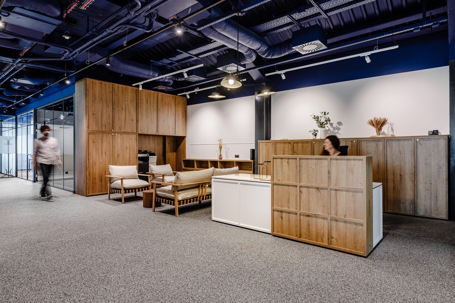 Nowoczesne biuro z drewnianymi akcentami: recepcja, meble do przechowywania i strefa relaksu. Ciemnoniebieski sufit z widoczną instalacją. Osoba za biurkiem i przechodzień.