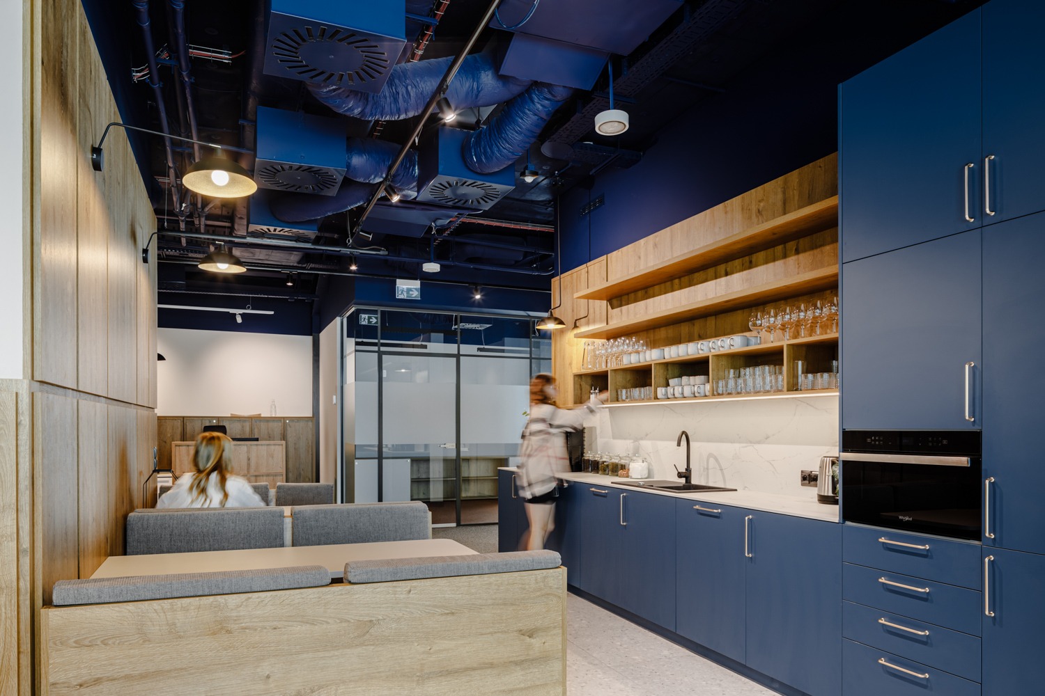 Nowoczesna aranżacja biurowej kuchni: niebieskie szafki, drewniane półki podświetlone LED, marmurowy blat, strefa relaksu z drewnianymi panelami i szarą tapicerką.