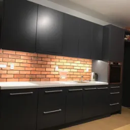 Nowoczesna kuchnia z czarnymi matowymi frontami, białym blatem i ceglaną ścianą. Widoczne oświetlenie podszafkowe i srebrne uchwyty.