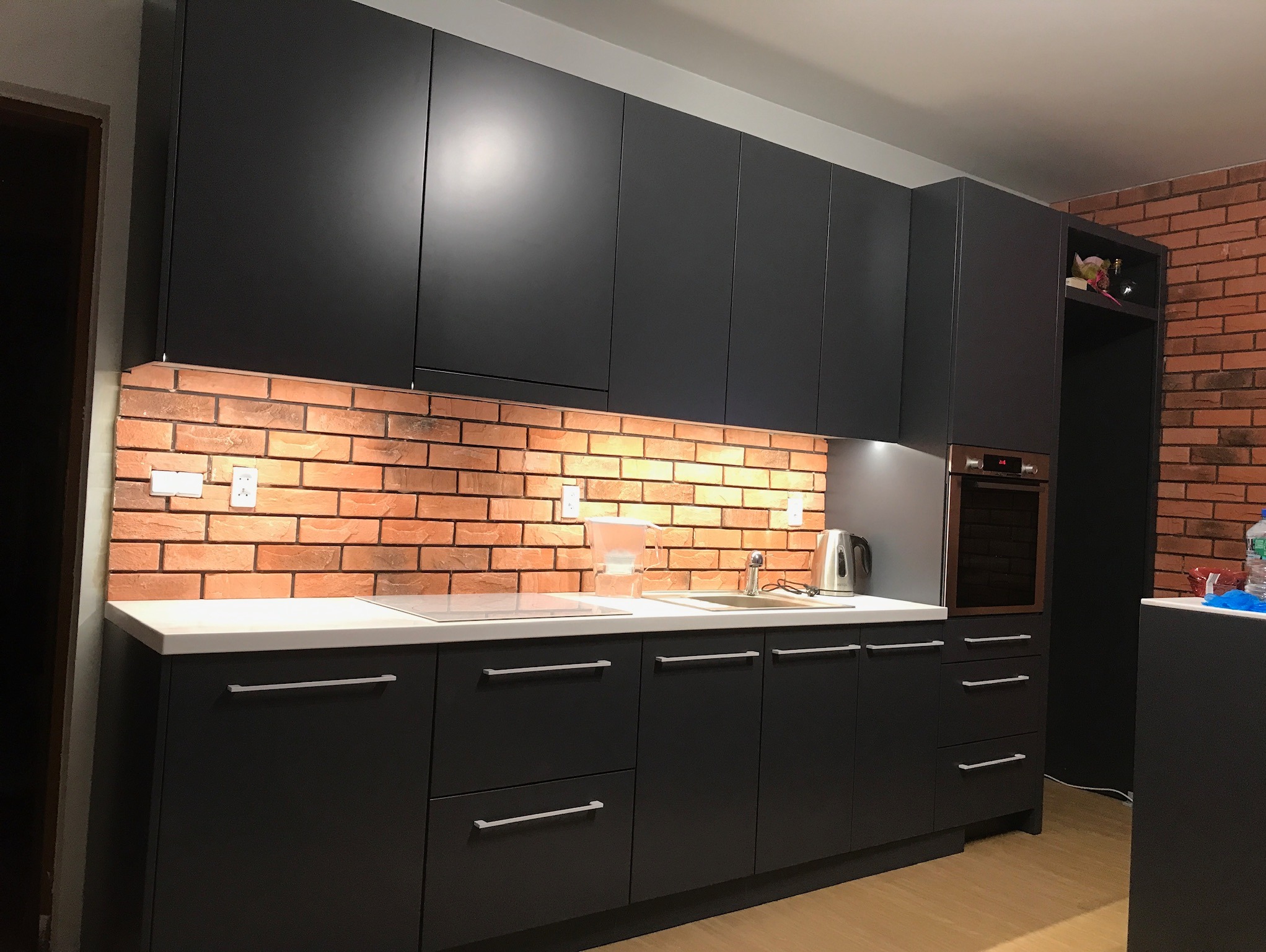 Nowoczesna kuchnia z czarnymi matowymi frontami, białym blatem i ceglaną ścianą. Widoczne oświetlenie podszafkowe i srebrne uchwyty.