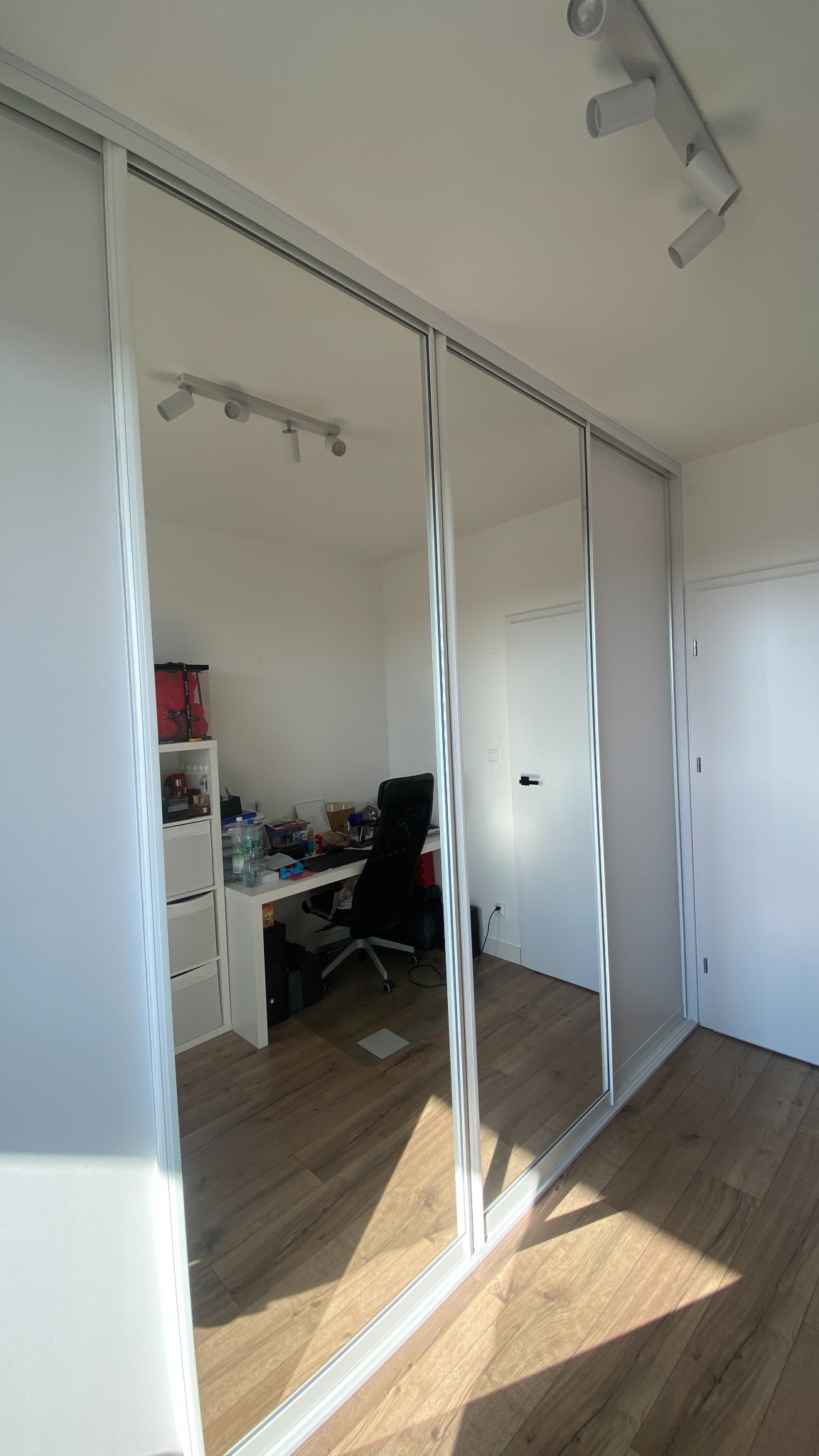 Biała szafa wnękowa z lustrzanymi drzwiami przesuwnymi, odbijająca pokój z biurkiem i krzesłem. Widoczne oświetlenie sufitowe i drewniana podłoga.