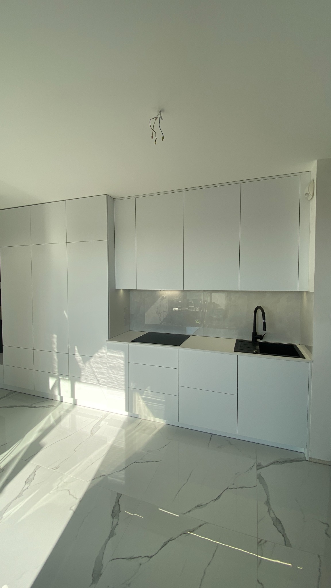 Nowoczesna, jasna kuchnia z białymi, minimalistycznymi szafkami i marmurową podłogą. Widoczne czarne elementy: zlew i płyta grzewcza. Na suficie wystające kable.