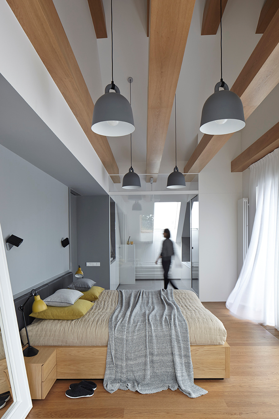 Nowoczesna sypialnia z drewnianymi belkami stropowymi i szarymi lampami wiszącymi. Minimalistyczny design, jasne kolory, osoba w tle przechodząca do łazienki.
