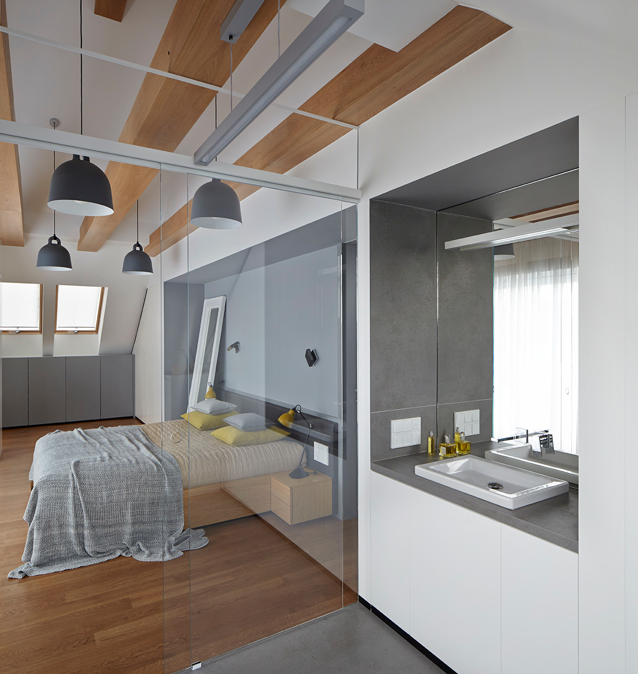 Nowoczesna sypialnia z łazienką oddzielona szklaną ścianą. Drewniane belki sufitowe, szare lampy i minimalistyczne meble. Widok na okna dachowe.