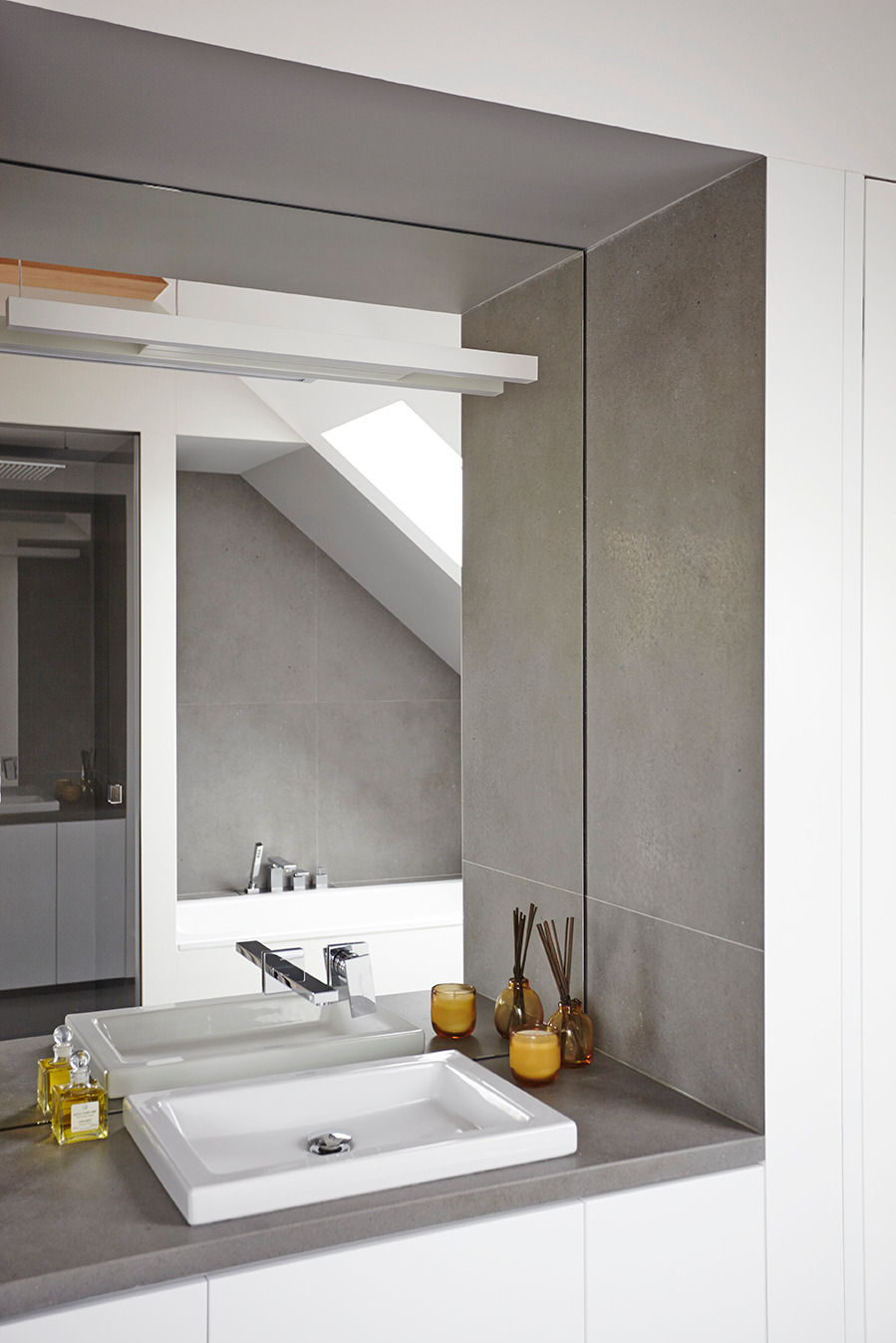 Nowoczesna łazienka z białą umywalką nablatową, chromowaną armaturą i szarymi płytkami. W lustrze odbija się wanna i okno dachowe. Minimalistyczny design.