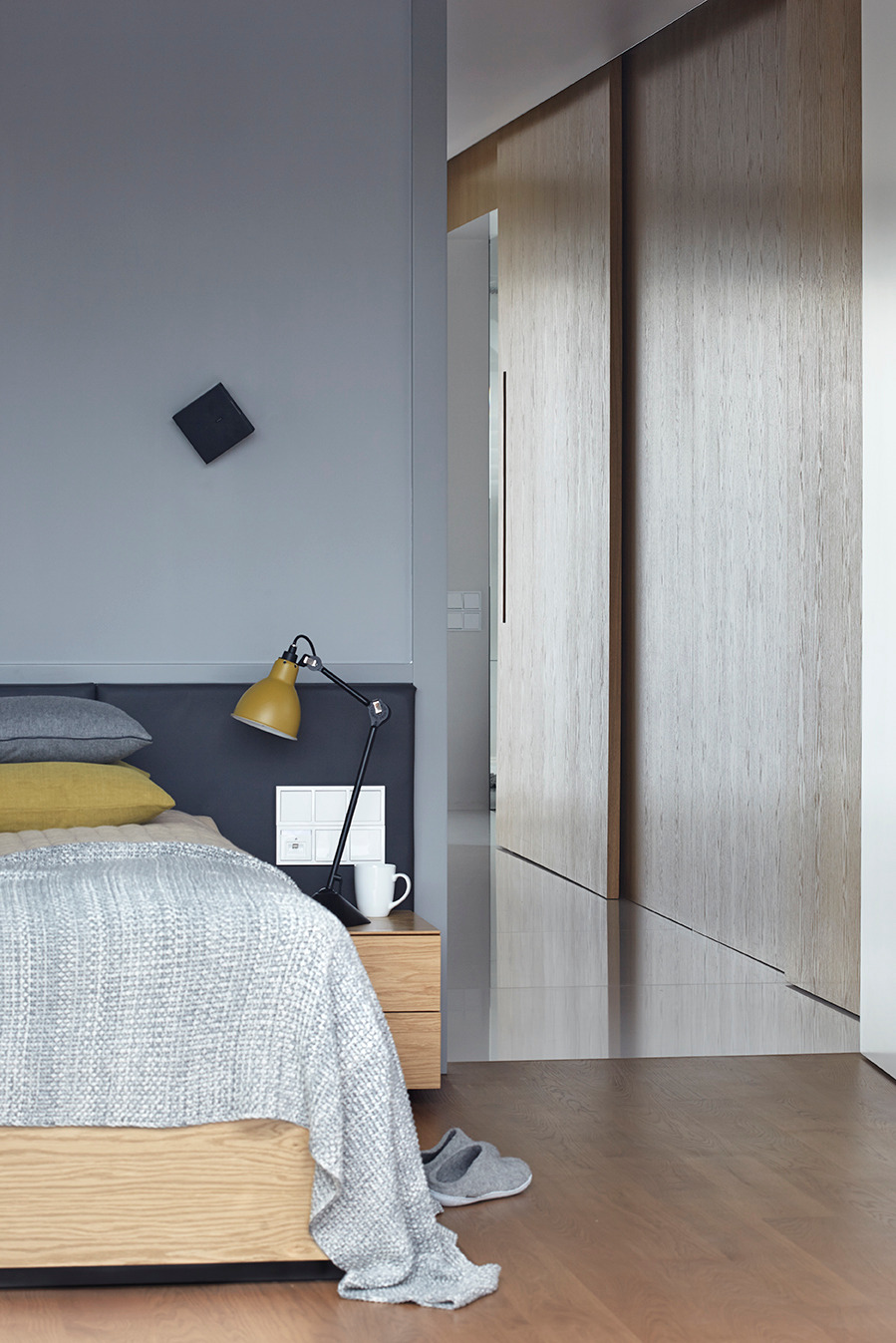 Nowoczesna sypialnia z przesuwnymi drzwiami szafy wnękowej z jasnego drewna. Na pierwszym planie łóżko z drewnianą ramą i szarą narzutą. Minimalistyczny design.