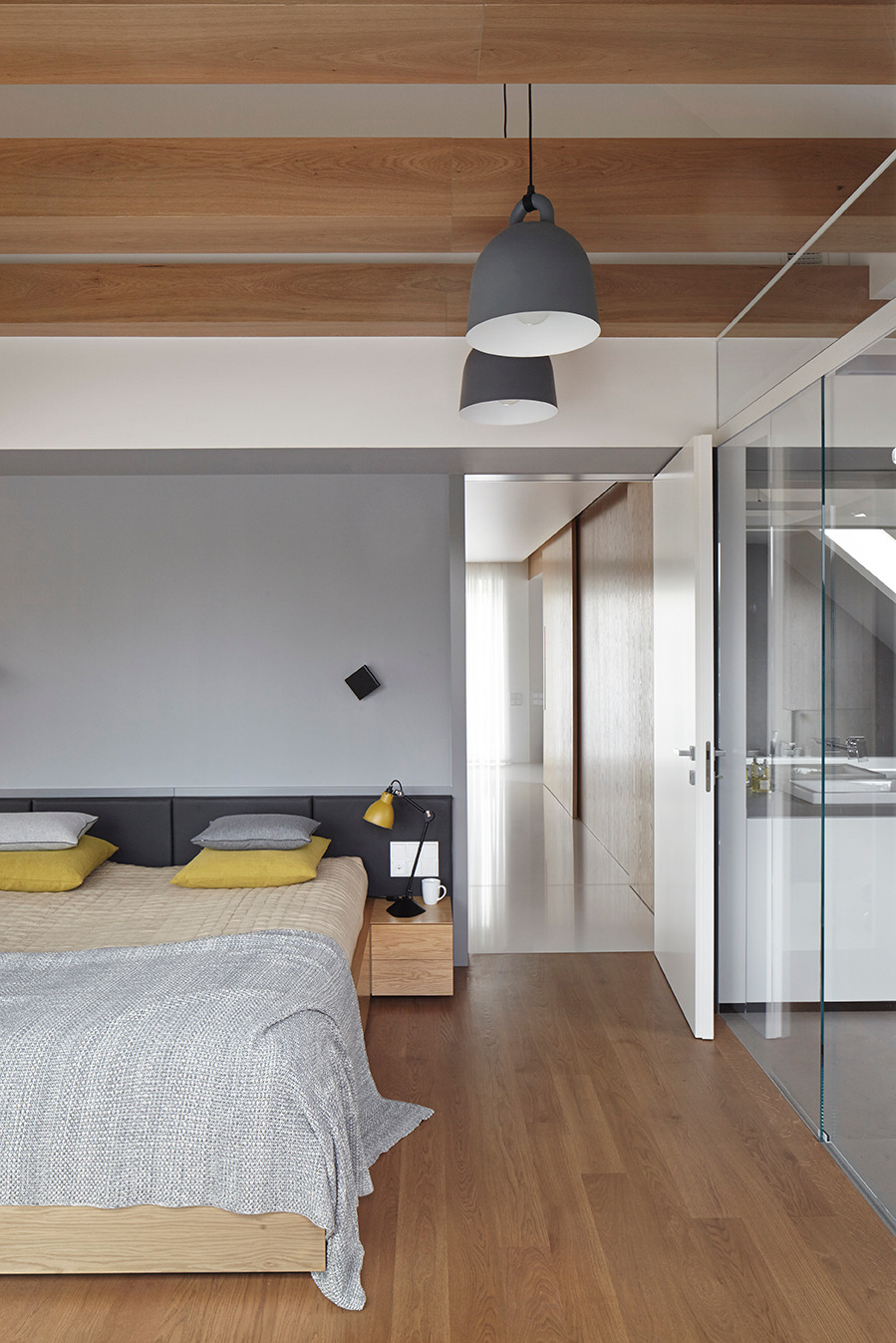 Nowoczesna sypialnia z drewnianą podłogą i belkami stropowymi, szara ściana, lampy wiszące i widok na łazienkę przez szklaną ścianę.