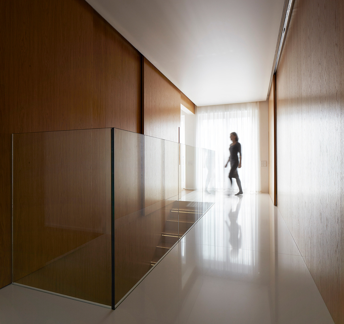Nowoczesny korytarz z drewnianą zabudową ścian, szklaną balustradą i polerowaną podłogą. W tle sylwetka kobiety przechodzącej przez jasne pomieszczenie.