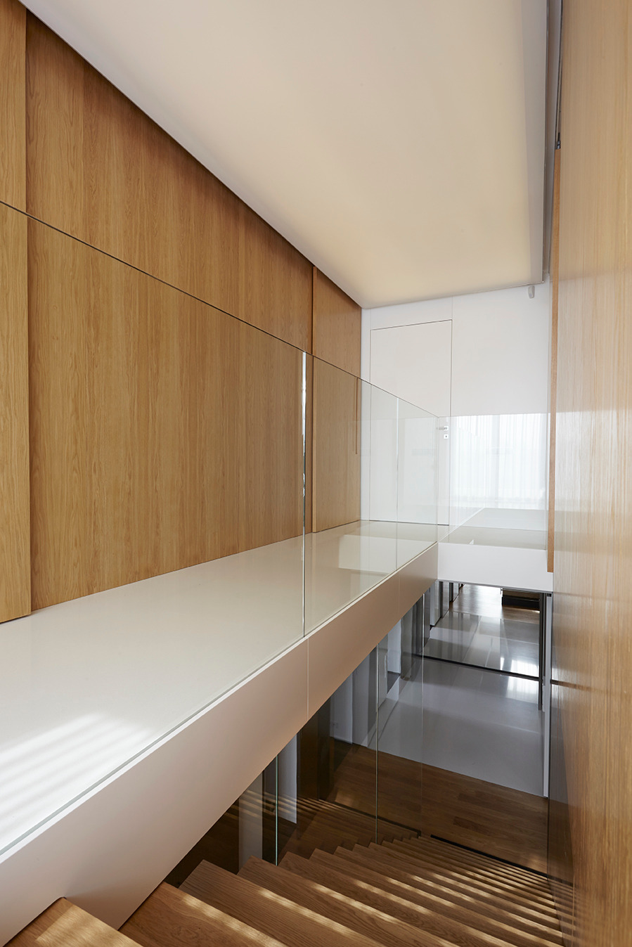 Nowoczesne schody z drewnianymi stopniami i szklaną balustradą, oświetlone naturalnym światłem, w minimalistycznym wnętrzu z drewnianą okładziną ścienną.