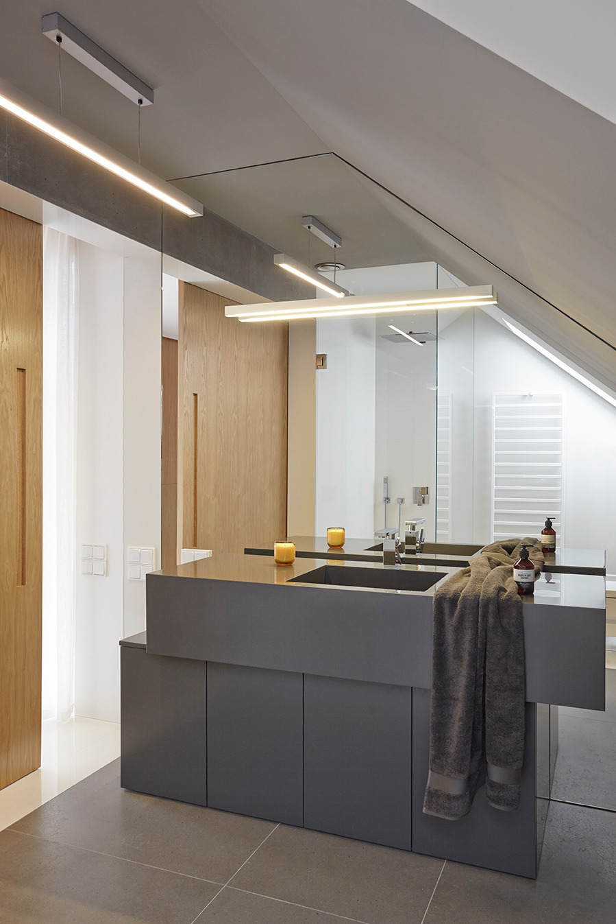 Nowoczesna łazienka na poddaszu z szarą szafką pod umywalkę, drewnianymi drzwiami i oświetleniem LED. Minimalistyczny design z ręcznikiem i akcesoriami.