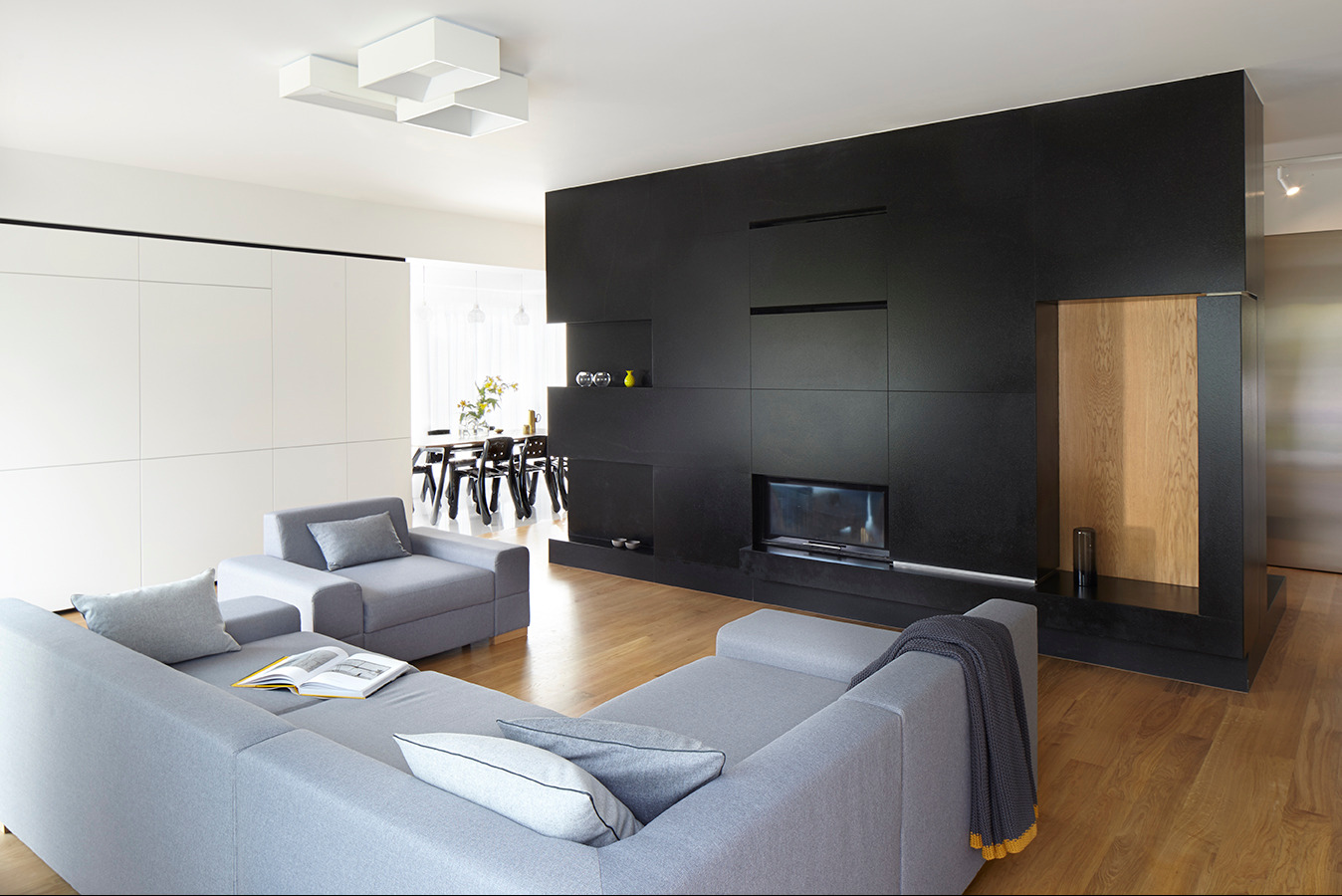 Nowoczesny salon z szarą sofą, czarną ścianą z wbudowanym kominkiem i białymi szafami w tle. Minimalistyczne oświetlenie i drewniana podłoga.