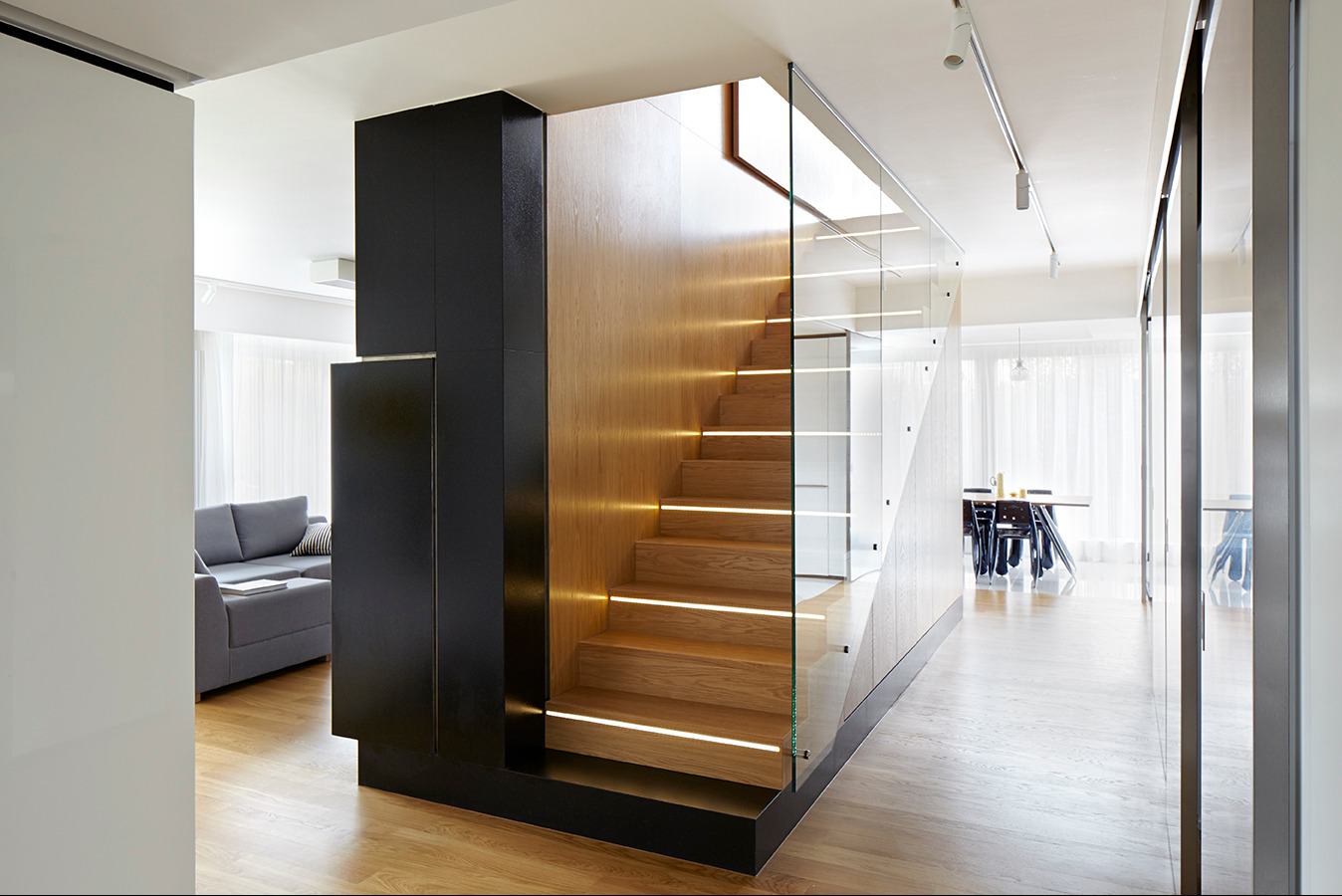 Nowoczesne schody z drewnianymi stopniami i podświetleniem LED, oddzielone szklaną balustradą, zintegrowane z czarną zabudową meblową w jasnym wnętrzu.