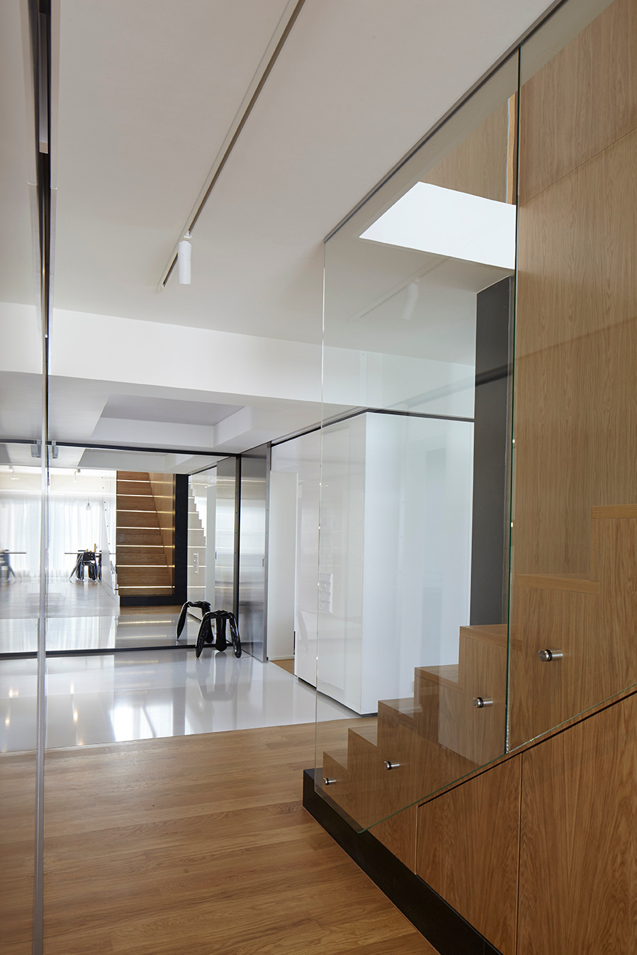 Nowoczesne schody z drewnianymi stopniami i szklaną balustradą, widoczne z perspektywy hallu z drewnianą podłogą. Minimalistyczny design, jasne wnętrze.