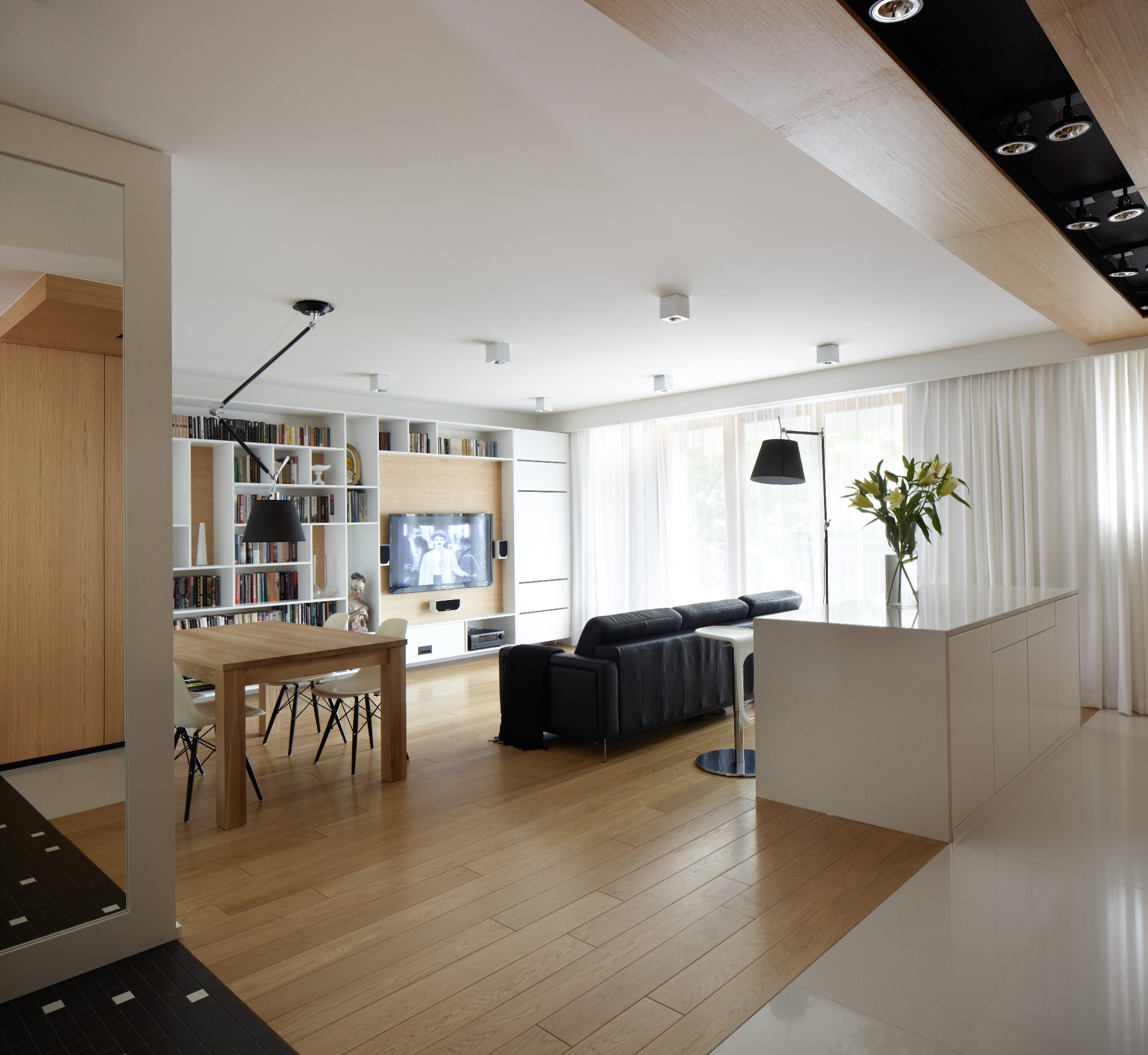 Jasne, przestronne wnętrze z drewnianą podłogą, białymi meblami i czarną sofą. Widoczny stół z krzesłami, regał z książkami i telewizor. Minimalistyczny design.