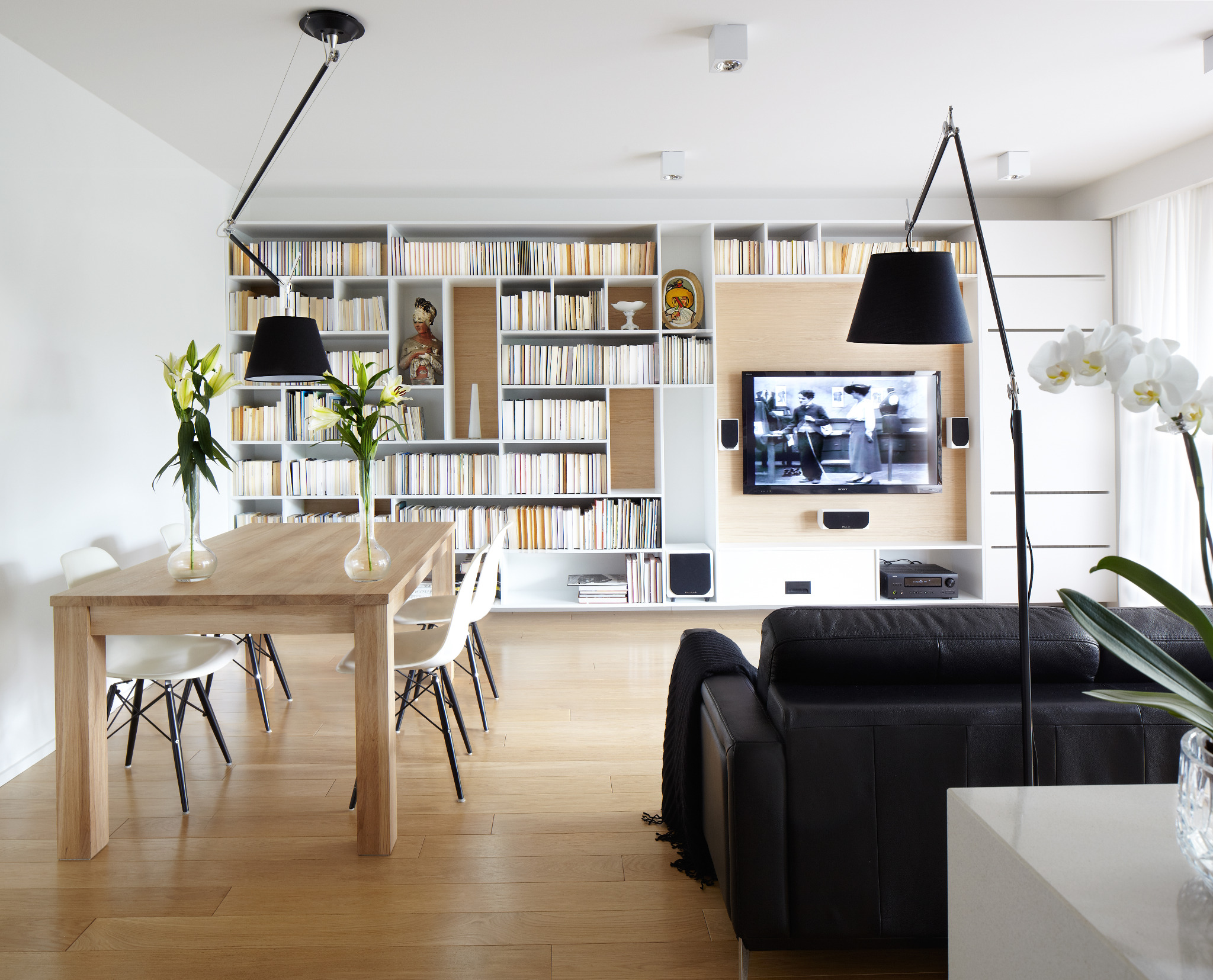 Jasne wnętrze: stół z kwiatami, regał z książkami, TV i czarna sofa. Minimalistyczny design, drewniana podłoga, lampy podłogowe. Aranżacja salonu z jadalnią.