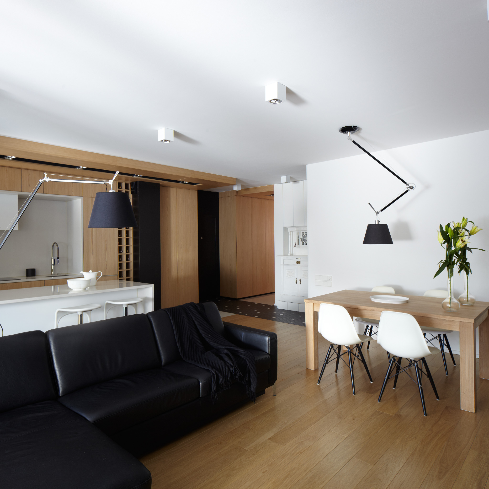 Nowoczesne wnętrze: czarna skórzana sofa narożna z narzutą, drewniany stół z białymi krzesłami, drewniana zabudowa kuchenna i podłoga, lampy designerskie.
