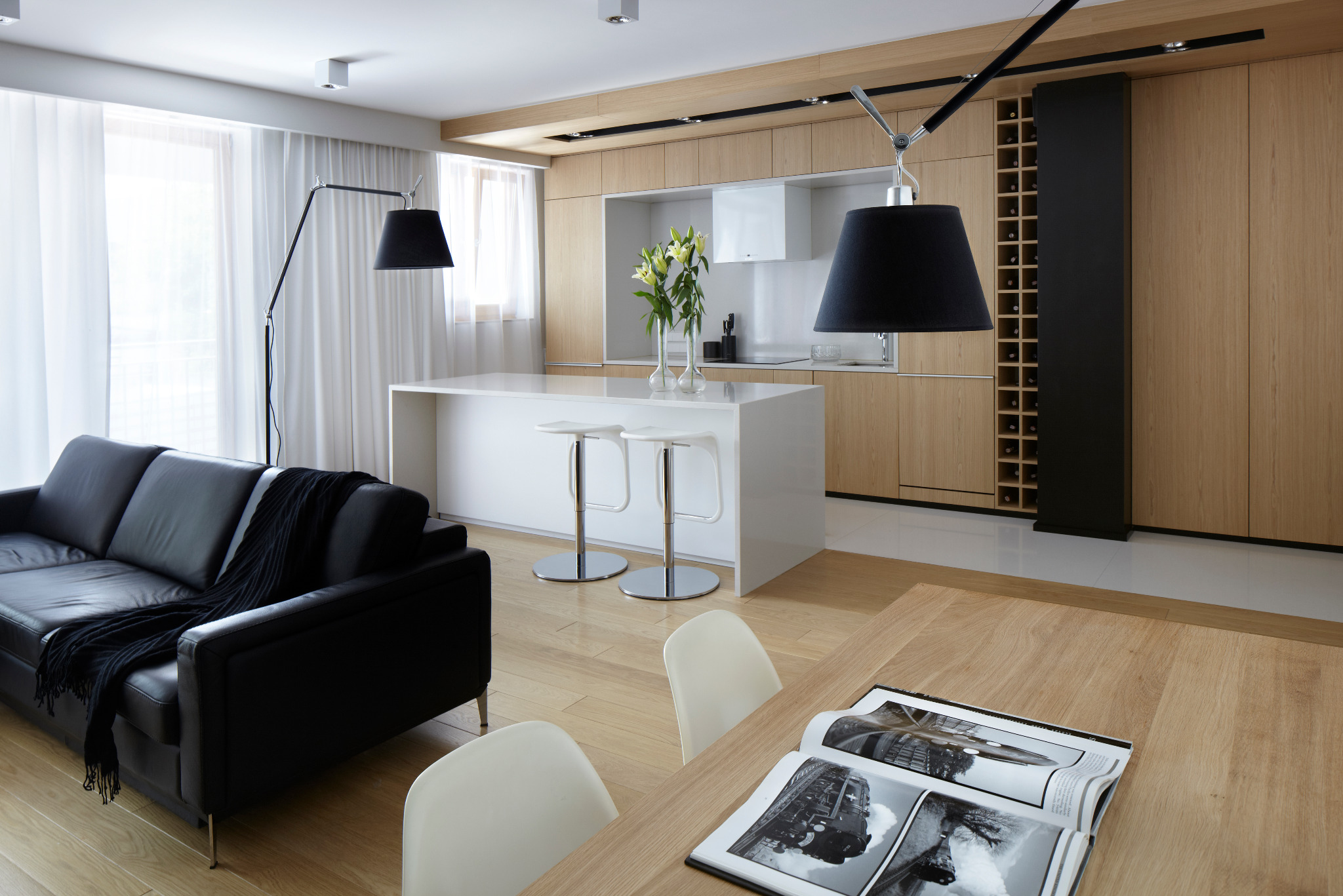 Nowoczesne wnętrze: drewniana zabudowa kuchenna z wyspą, czarna sofa i lampy. Na stole album z czarno-białymi zdjęciami. Minimalistyczny design, jasne kolory.