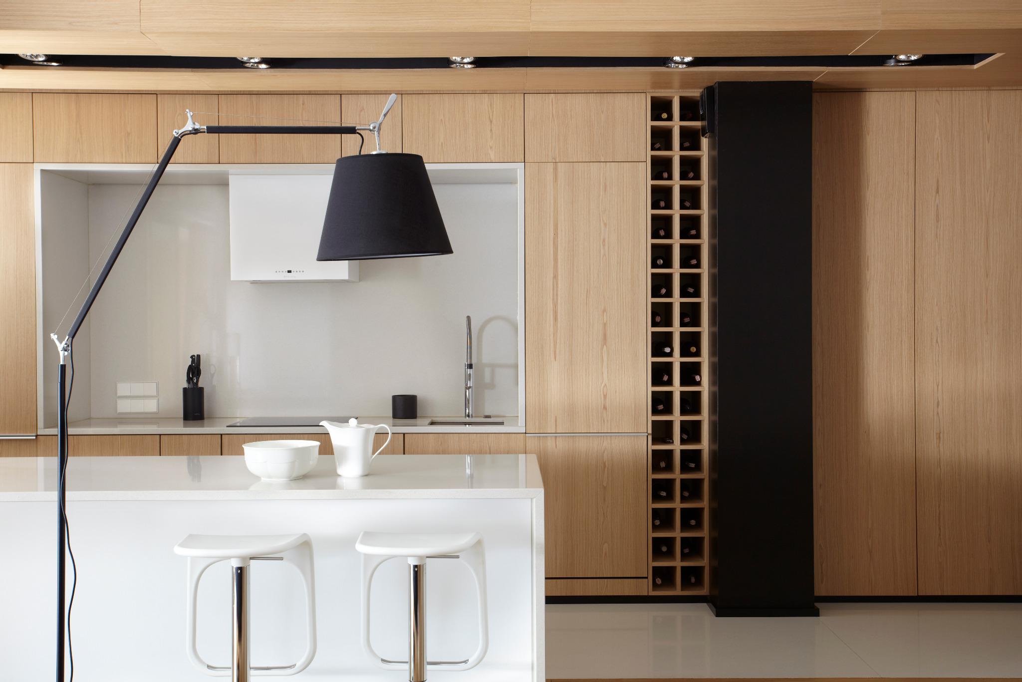 Nowoczesna kuchnia z drewnianymi frontami, wyspą z białym blatem i czarnym regałem na wino. Minimalistyczny design, oświetlenie punktowe i lampa nad wyspą.