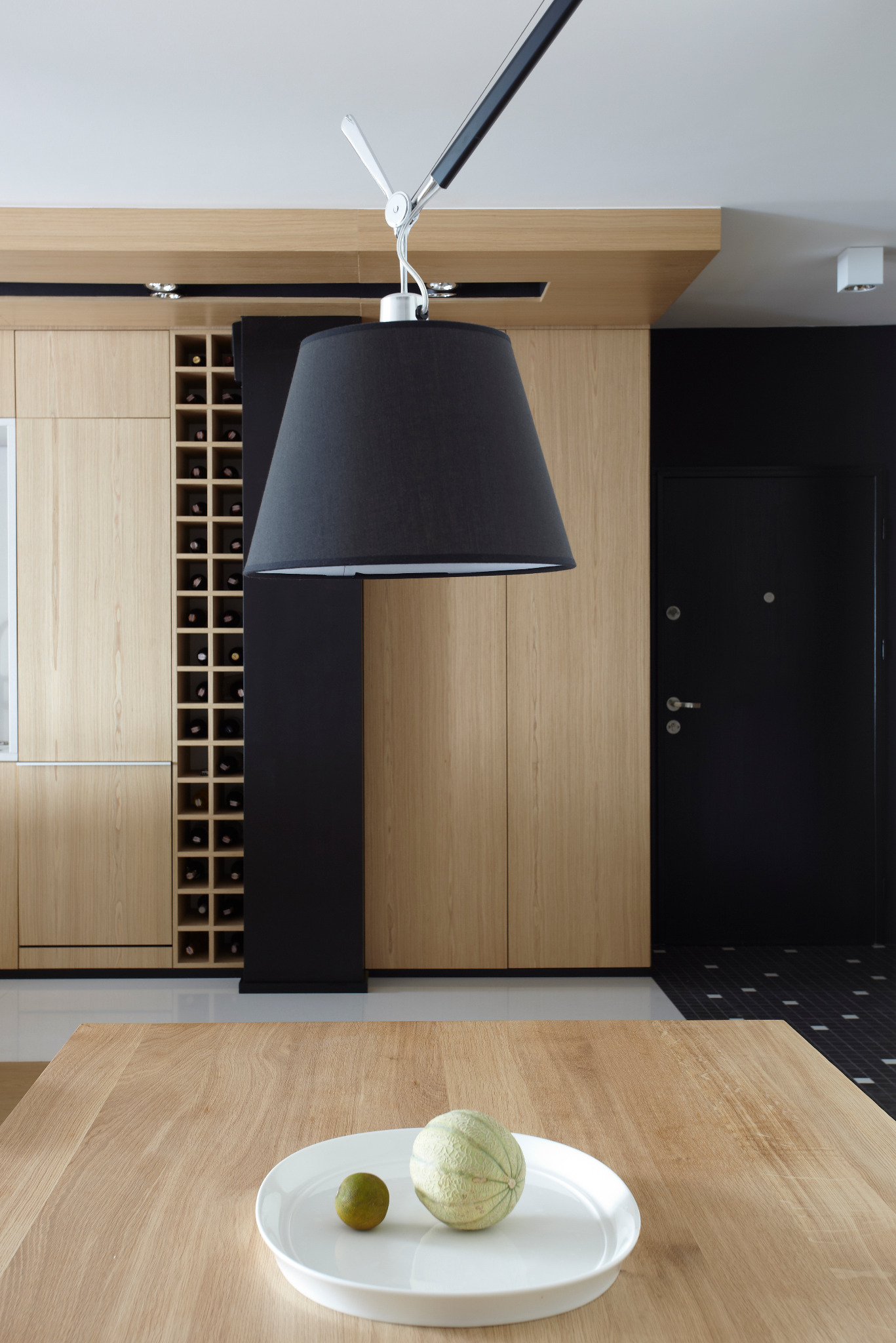 Nowoczesna zabudowa kuchenna z jasnego drewna z regałem na wino, czarnym akcentem i lampą. Na pierwszym planie stół z owocami na białym talerzu.