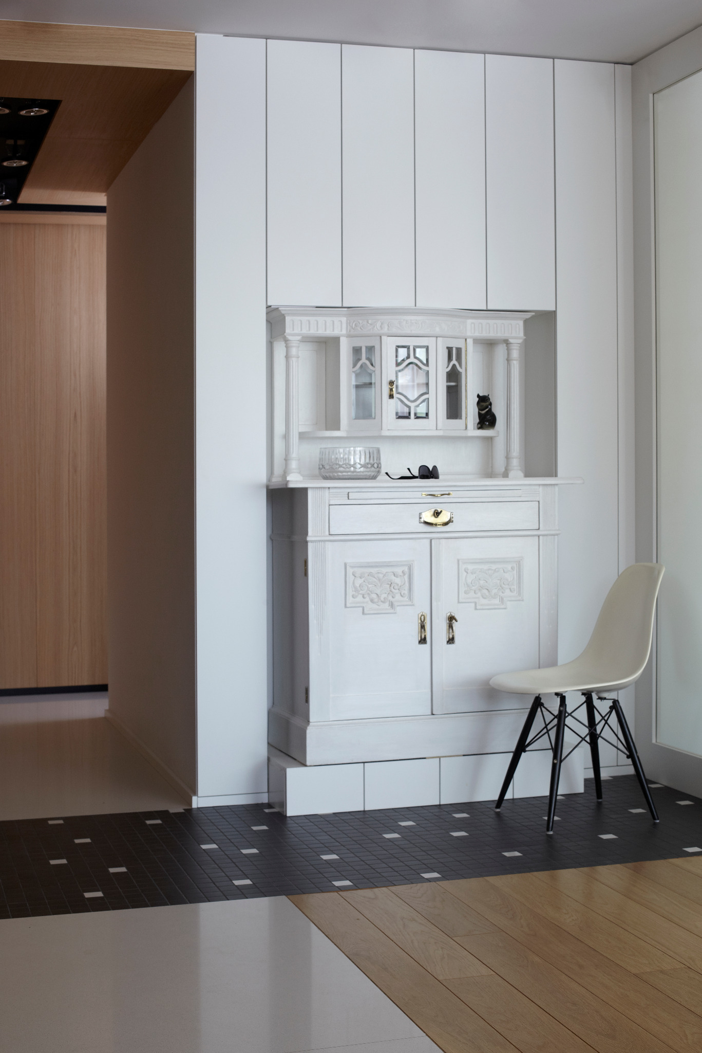 Biała, stylizowana komoda z nadstawką wkomponowana w ścianę z boazerią. Na blacie kryształowa misa i okulary. Obok krzesło na tle podłogi z mozaiką i drewnem.