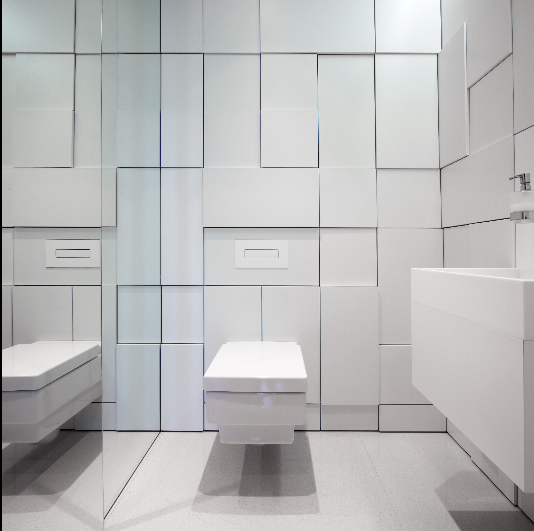 Minimalistyczna, biała łazienka z geometryczną ścianą z płytek, podwieszaną toaletą i umywalką. Nowoczesny design z subtelnym oświetleniem.