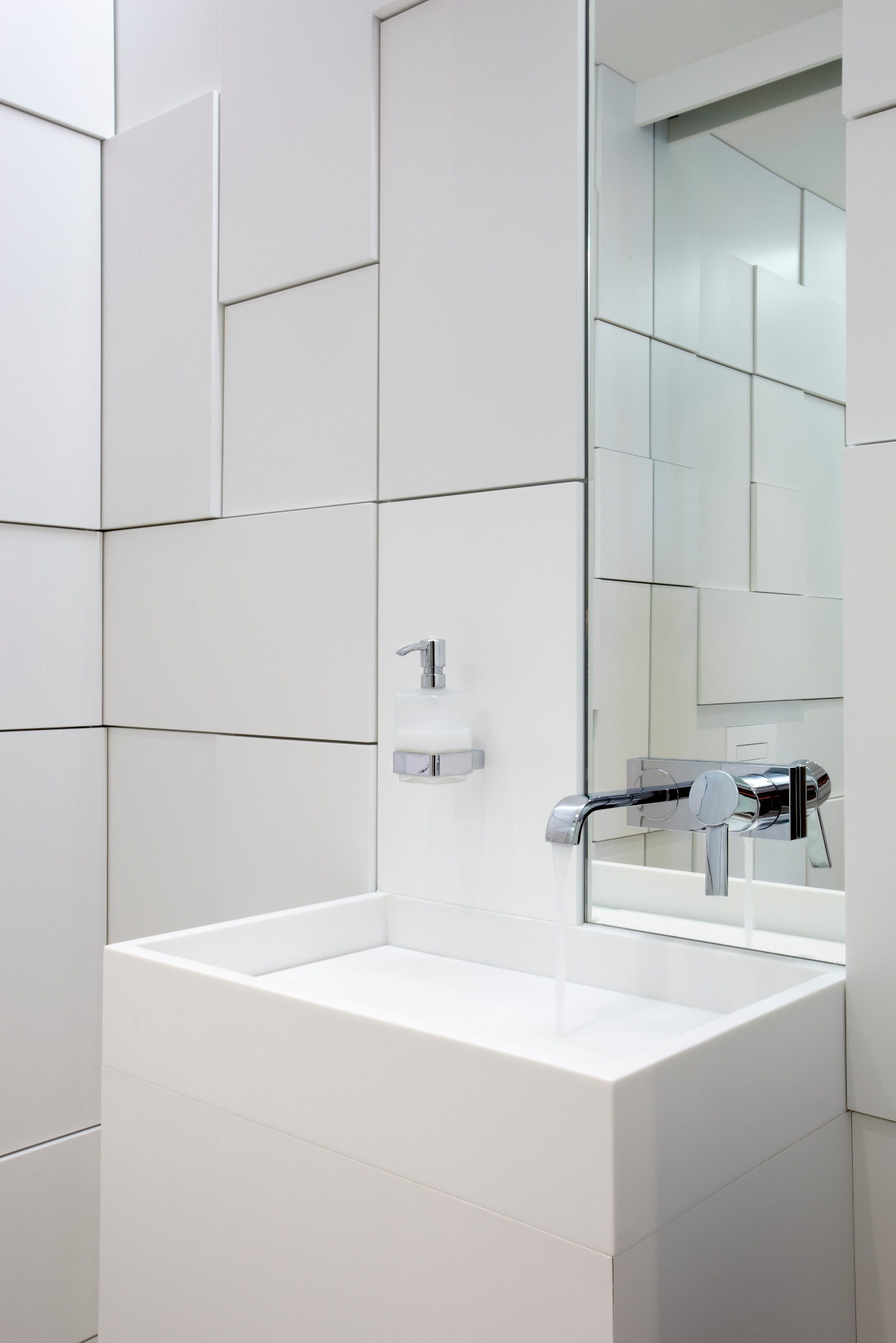 Minimalistyczna, biała łazienka z geometryczną ścianą, kwadratową umywalką i chromowaną baterią, z której leje się woda. Nowoczesny design.