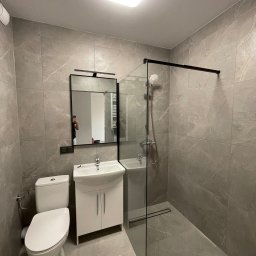 Renovatech Roman Ocheretnyi - Szara łazienka z kabiną prysznicową typu walk-in, białą umywalką z szafką i toaletą. Na ścianach i podłodze płytki imitujące marmur.