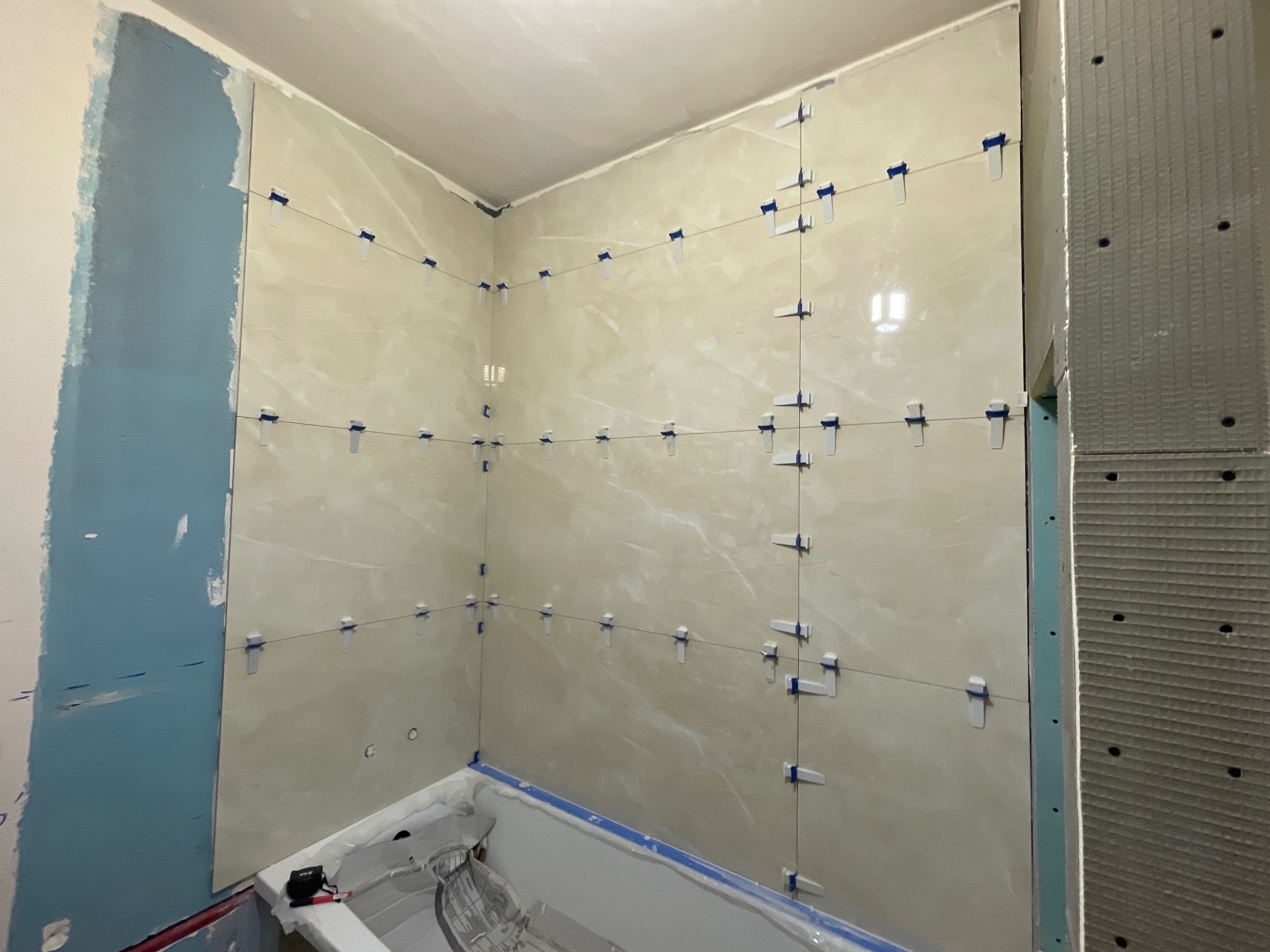 Łazienka w trakcie remontu: beżowe płytki imitujące marmur, niebieskie elementy zabezpieczające, widoczna wanna i fragment ściany z płytą kartonowo-gipsową.