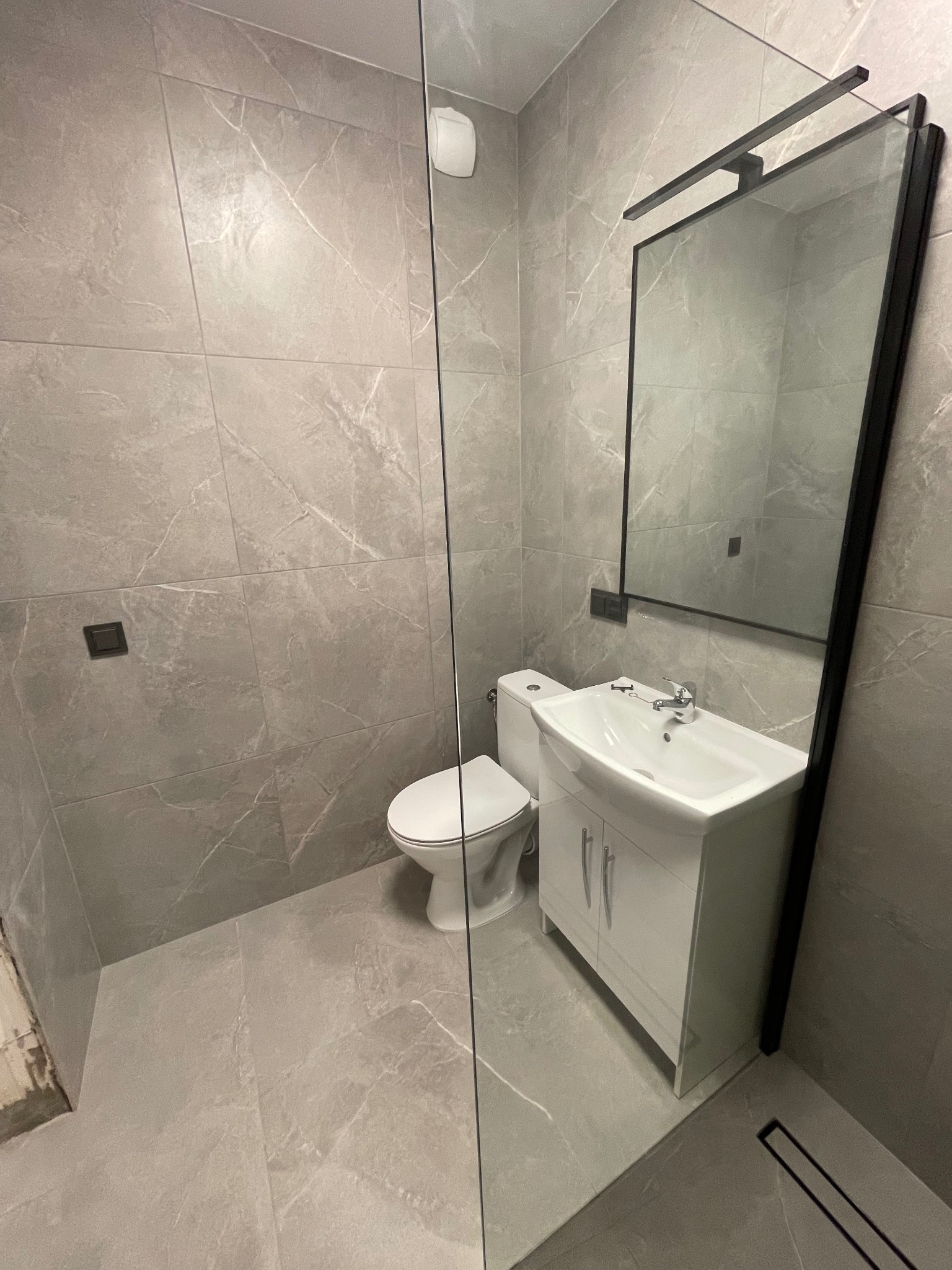 Nowoczesna łazienka w szarościach: płytki imitujące marmur, biała umywalka z szafką, toaleta i minimalistyczne lustro w czarnej ramie. Kabina prysznicowa ze szklaną ścianką.