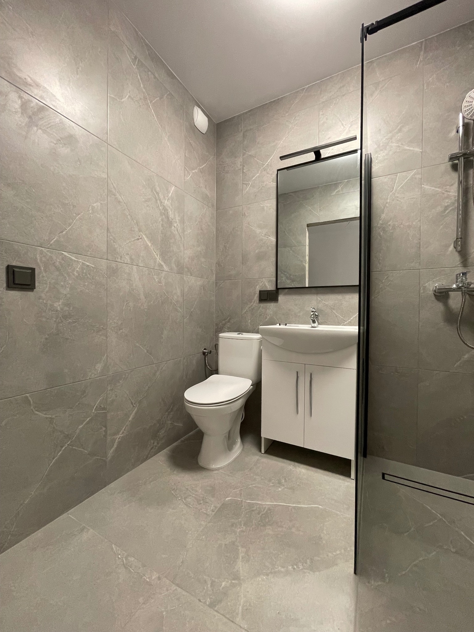 Wykończona łazienka w szarościach: płytki imitujące marmur na ścianach i podłodze, biała szafka z umywalką i lustrem w czarnej ramie, toaleta i kabina prysznicowa.
