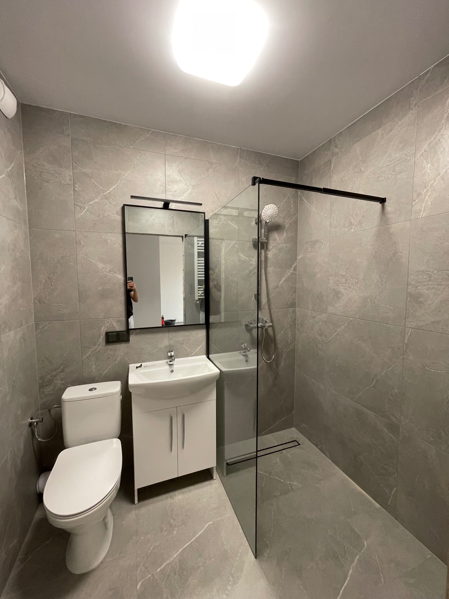 Szara łazienka z kabiną prysznicową typu walk-in, białą umywalką z szafką i toaletą. Na ścianach i podłodze płytki imitujące marmur.