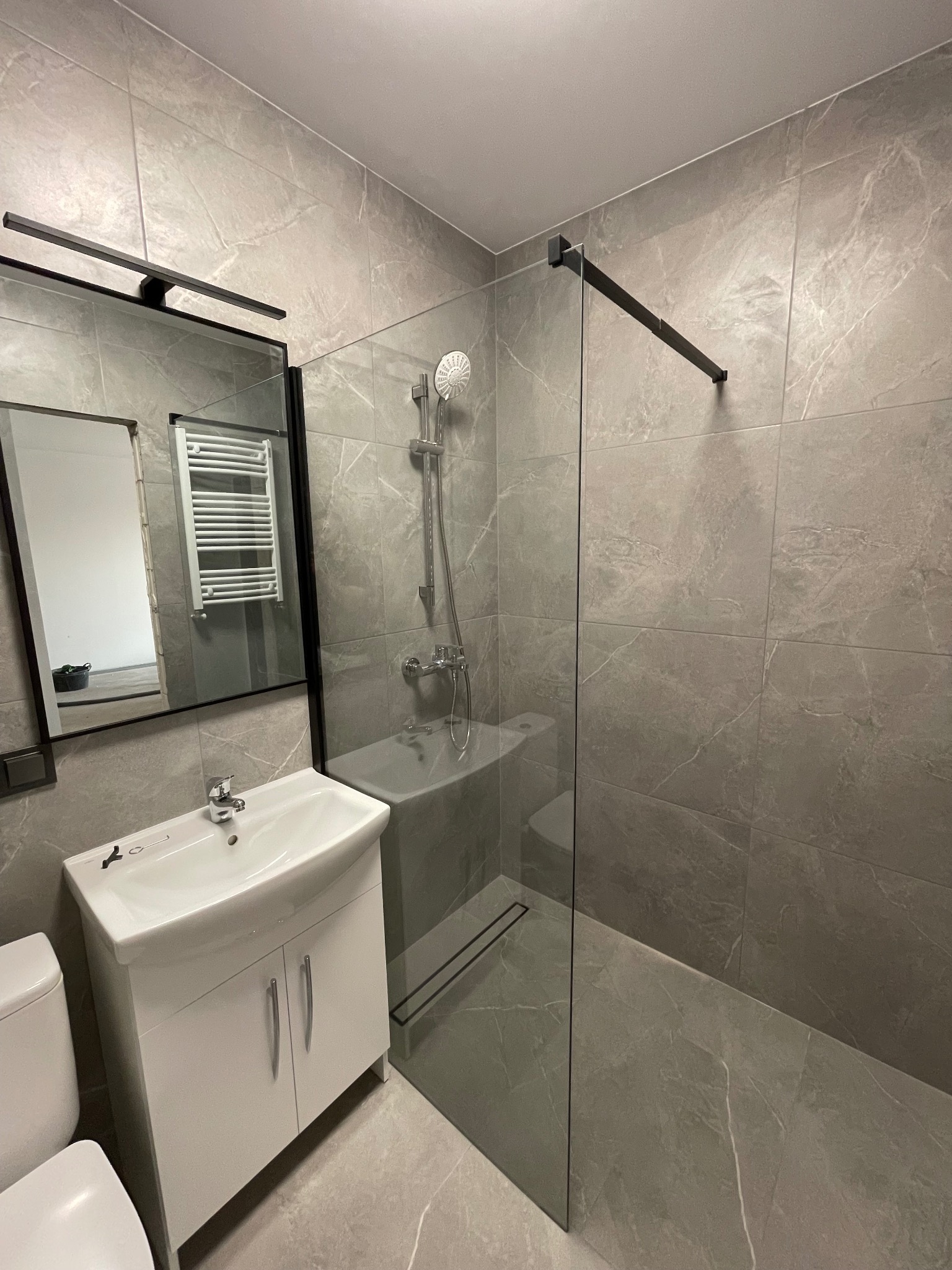 Nowoczesna łazienka w szarościach z kabiną prysznicową typu walk-in, białą szafką pod umywalką i lustrem z czarną ramą. Minimalistyczny design, stonowana kolorystyka.