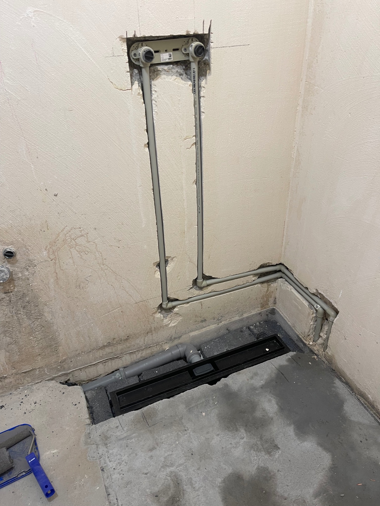 Szare rury wodne w ścianie i odpływ liniowy w surowej posadzce łazienki, widoczne ślady prac remontowych.