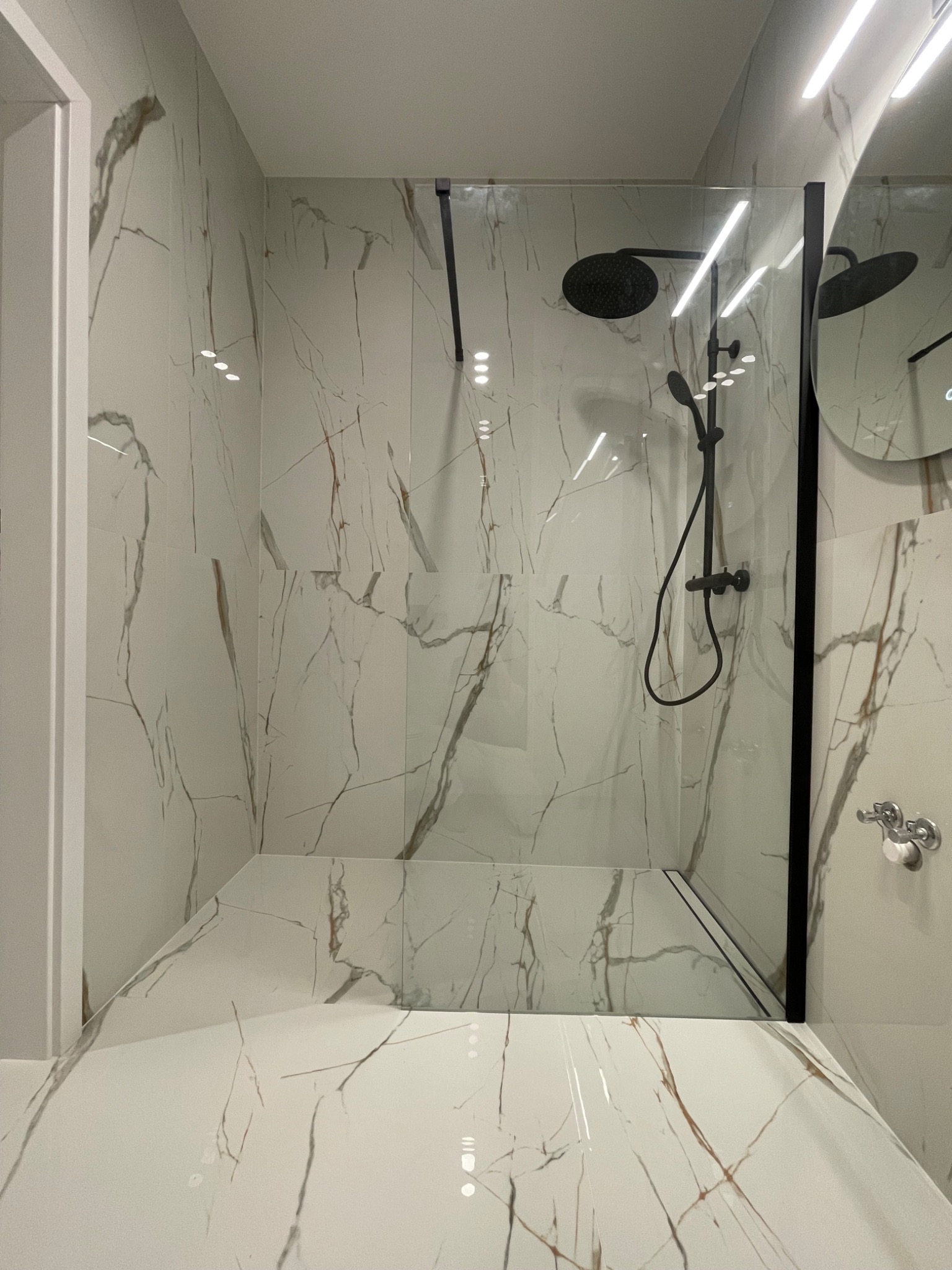 Nowoczesna kabina prysznicowa typu walk-in z czarną armaturą i białymi płytkami imitującymi marmur z brązowymi żyłkami, tworząca elegancką przestrzeń kąpielową.