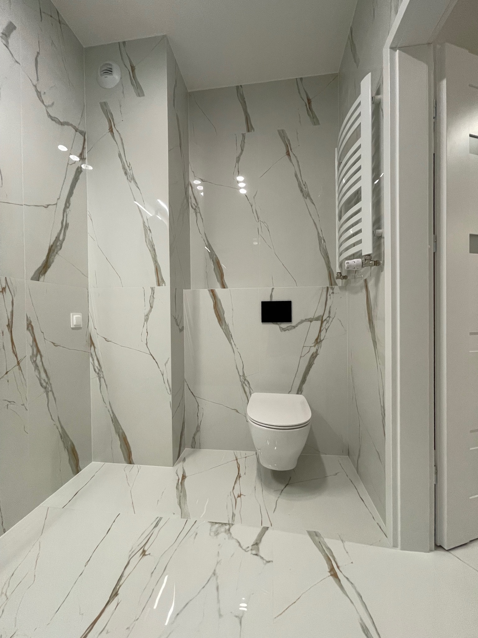 Biała łazienka z marmurowymi płytkami, wiszącą toaletą i grzejnikiem drabinkowym. Nowoczesne wykończenie z akcentami imitacji marmuru na ścianach i podłodze.