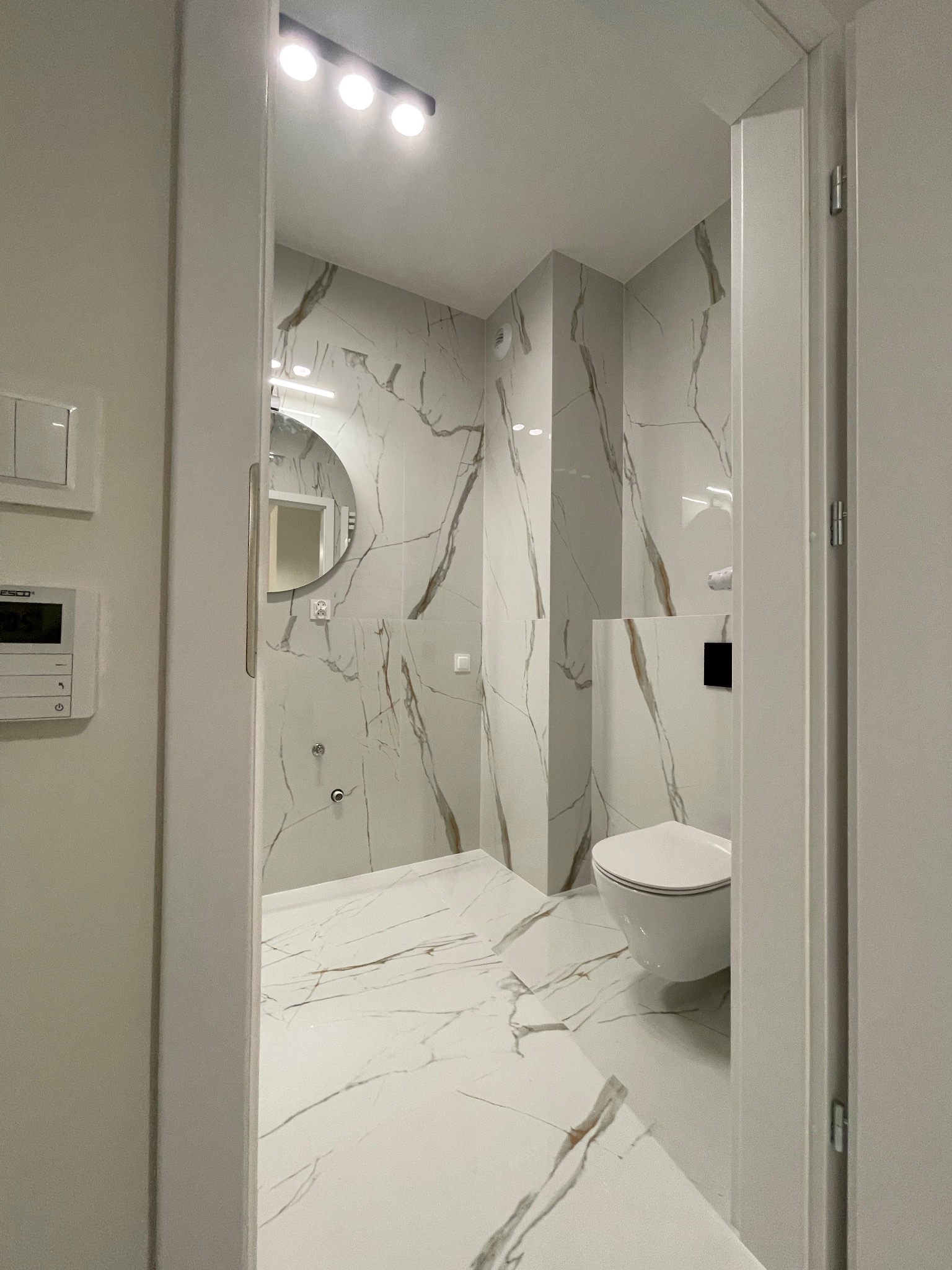 Elegancka łazienka w Warszawie z białymi płytkami imitującymi marmur, okrągłym lustrem i podwieszaną toaletą, widoczna zza uchylonych drzwi.